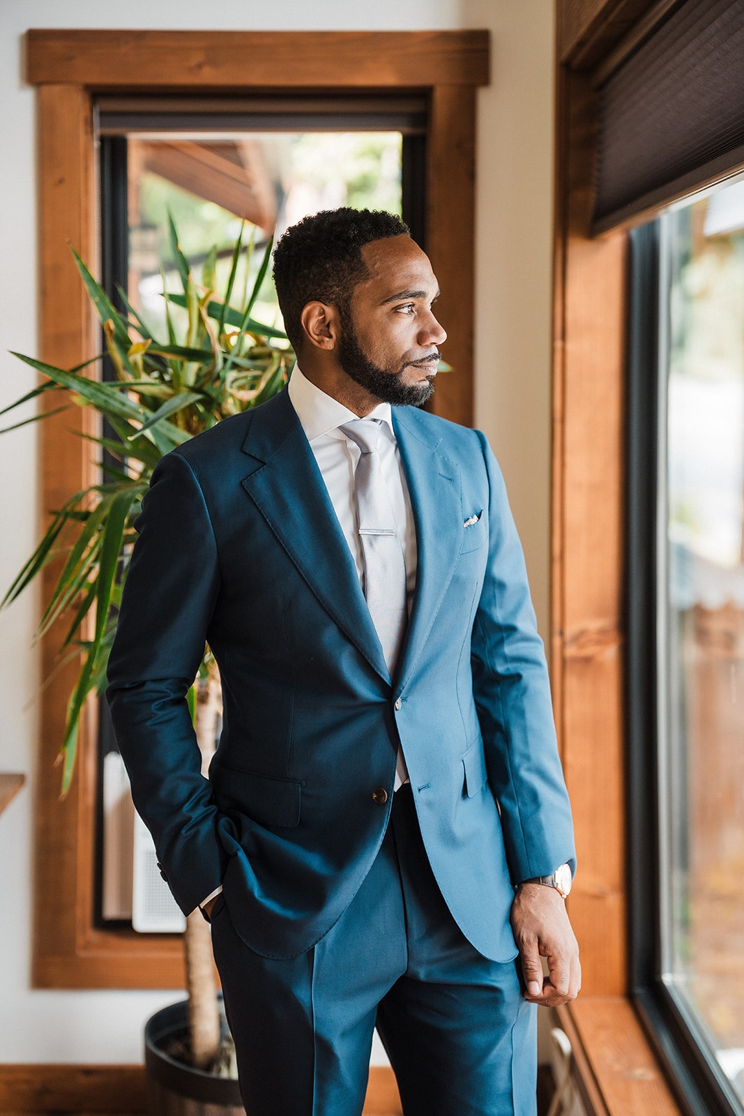 Groom in blue suit stands next to a window in his elopement cabin in Washington.