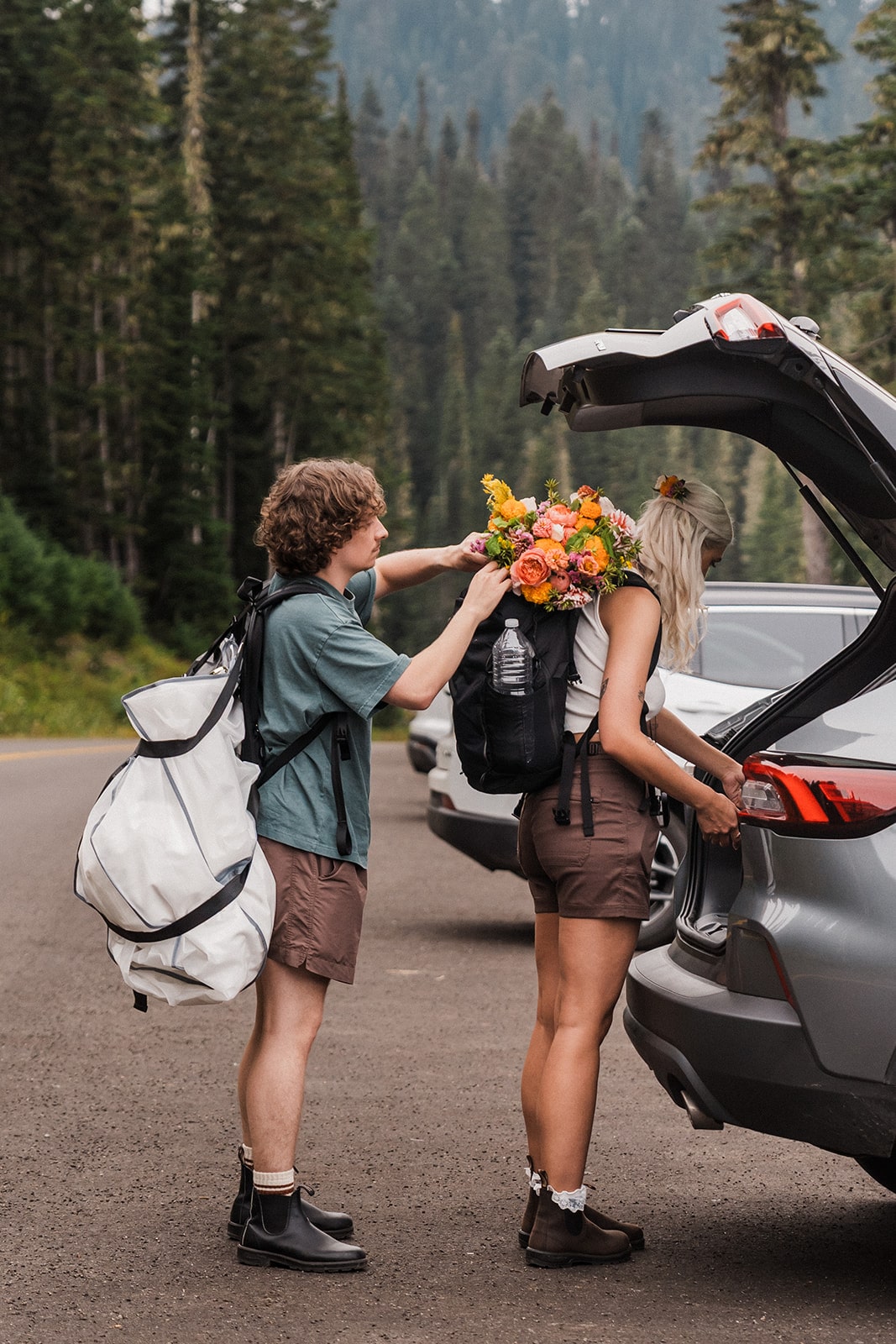Man clips elopement flowers to woman's hiking backpack as they prepare for their Washington elopement adventure.
