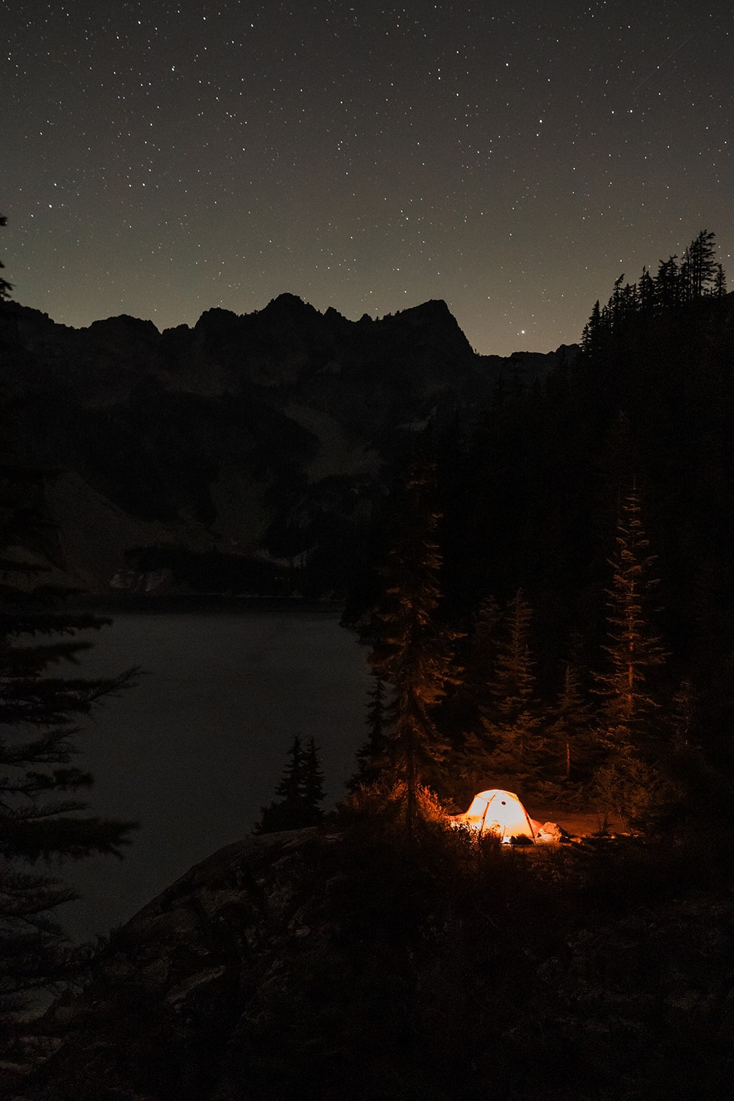 Tent lit with a lantern perches on the side of a mountain lake under a black starry sky.