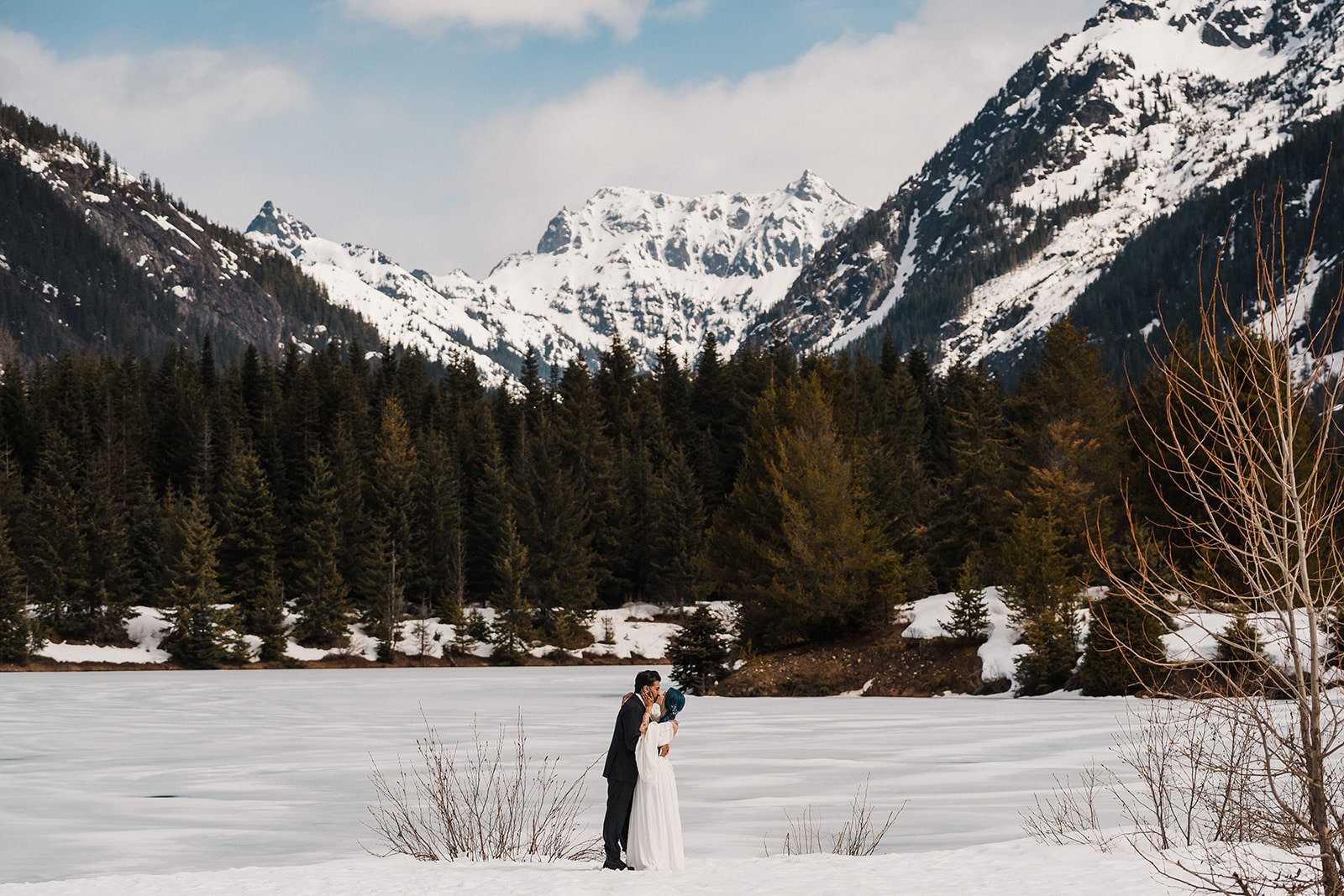 Bride and groom kiss next to a snowy Gold Creek Pond with snow-capped mountains in the background. 