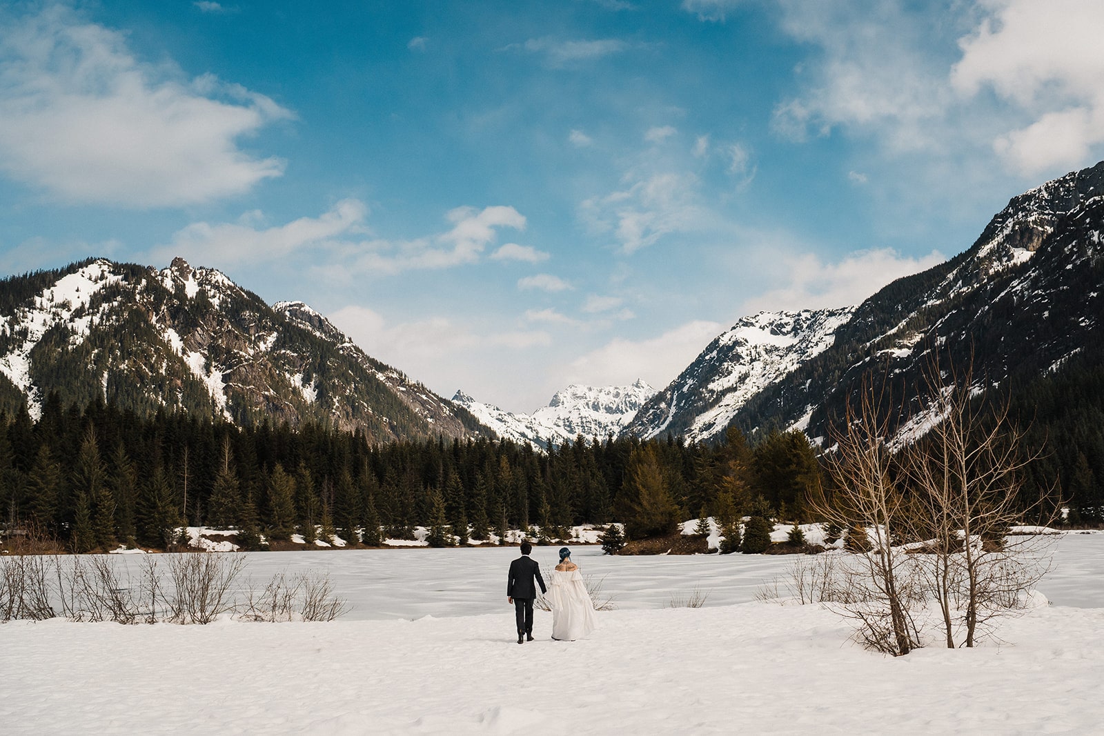 Bride and groom hold hands as they walk towards Gold Creek Pond in the snow. 