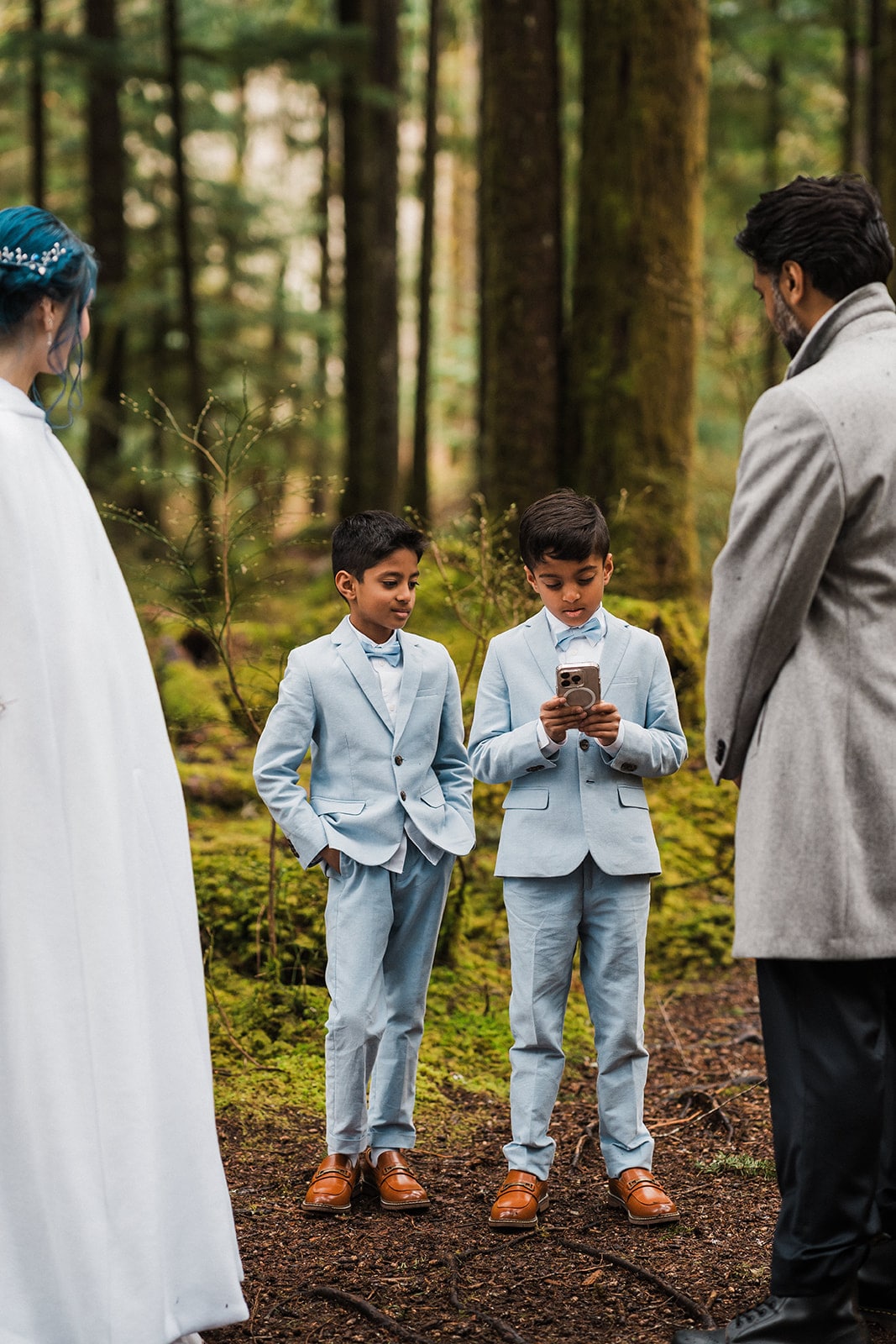 Two boys in blue suits read from an iPhone to the bride and groom during their unique elopement ceremony in the forest. 