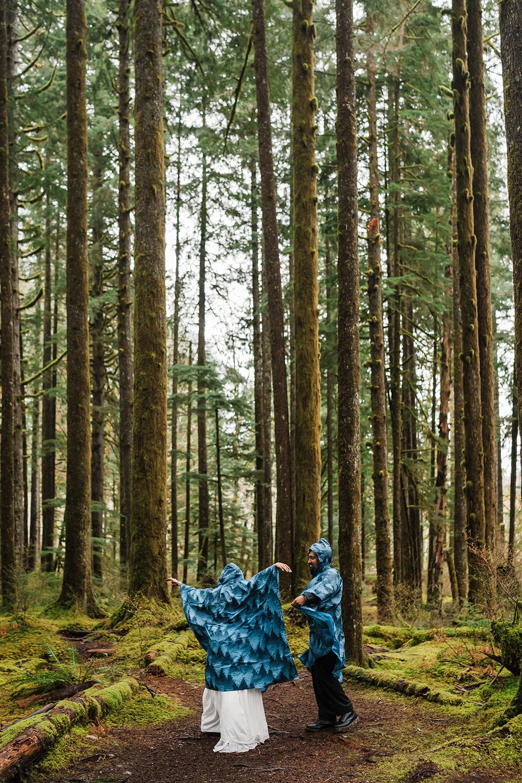 Bride and groom dance in the forest while wearing blue ponchos. 