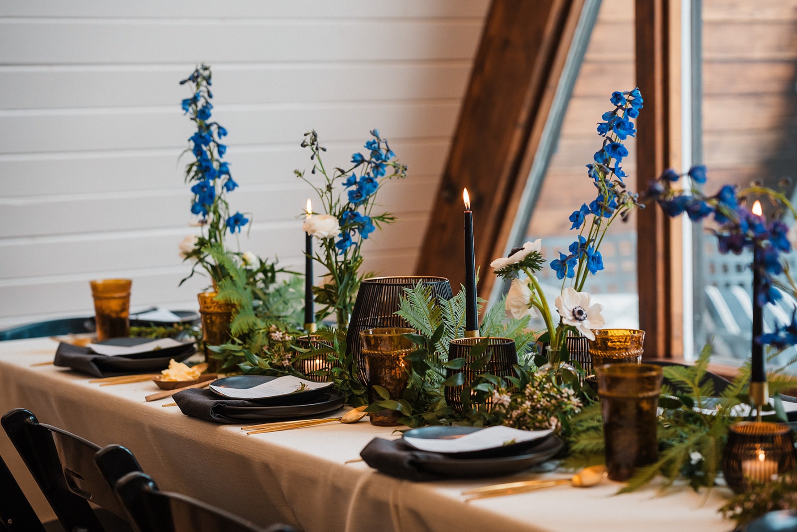 Blue flowers line the middle of a table with black taper candles and plates. 