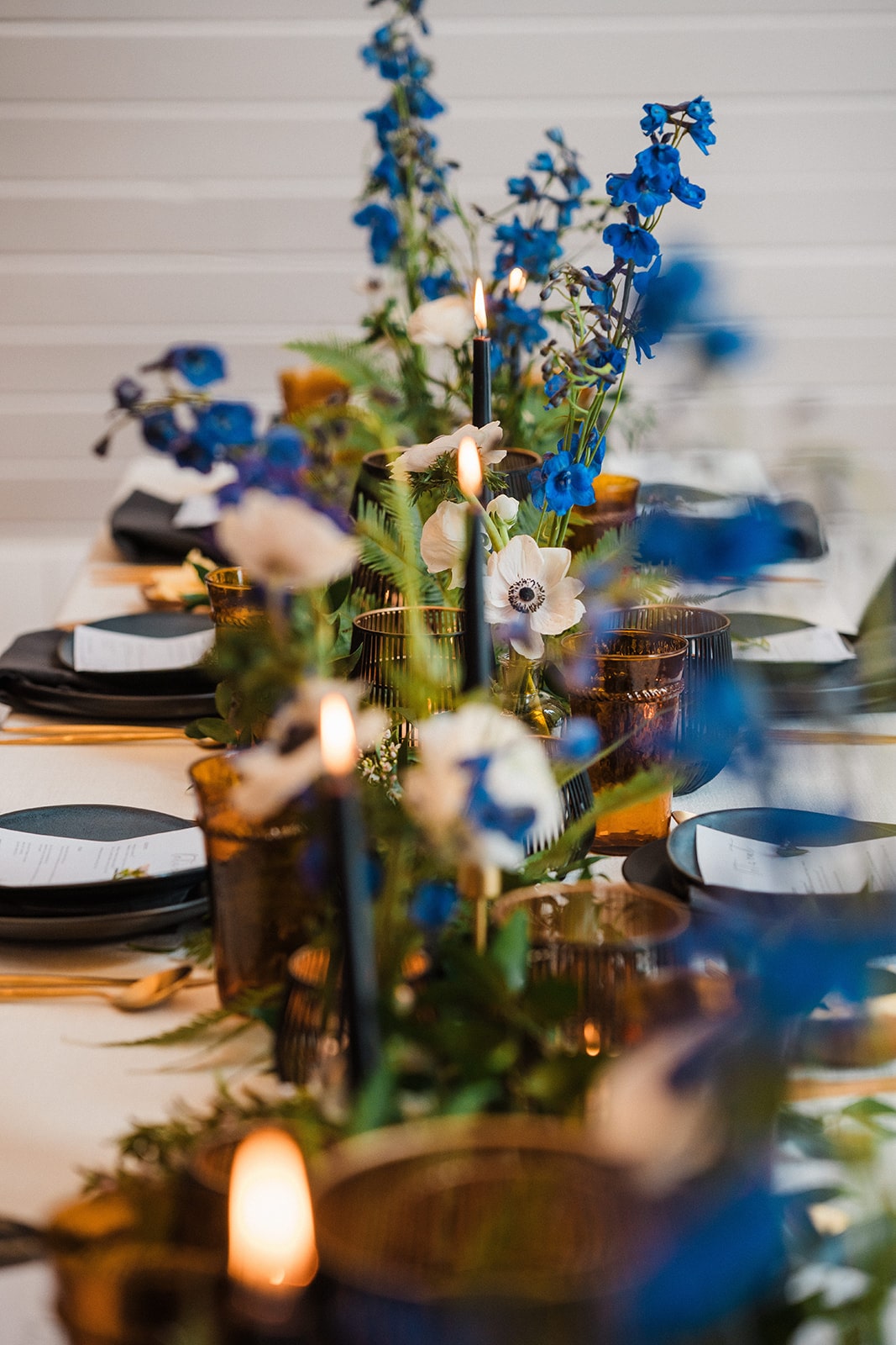 Blue flowers line the middle of a wedding reception dinner table with black taper candles and black plates. 