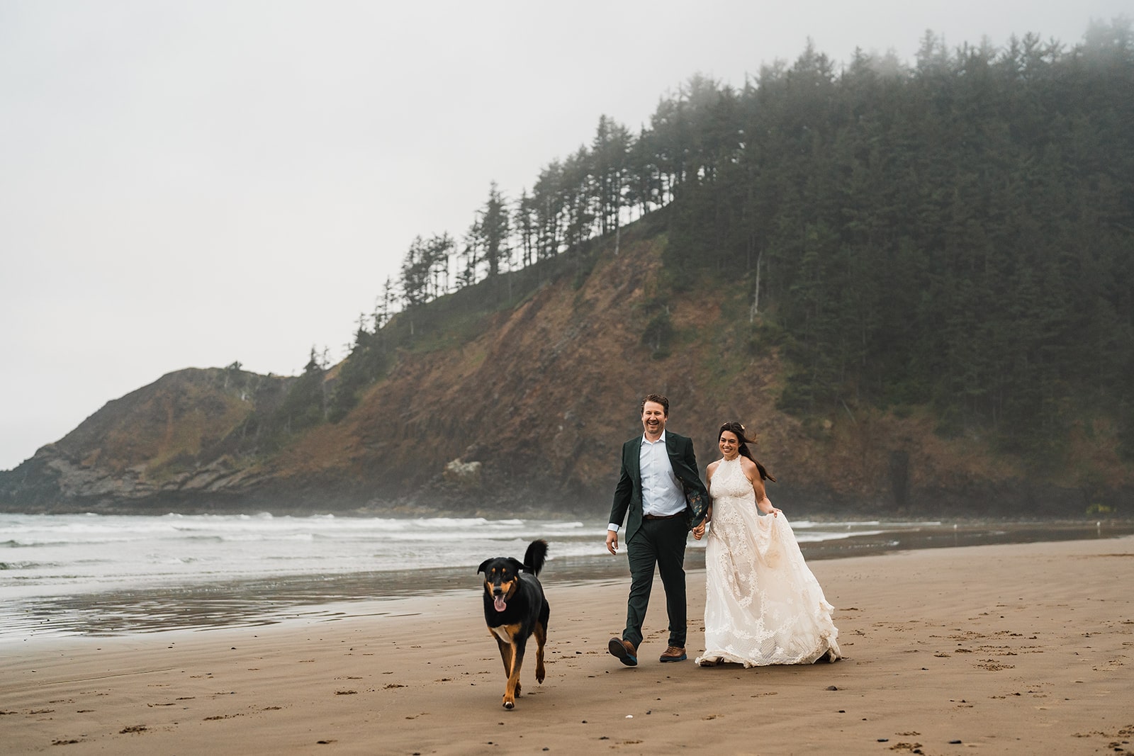 Bride and groom walk on a beach while their dog runs in front of them during their elopement.