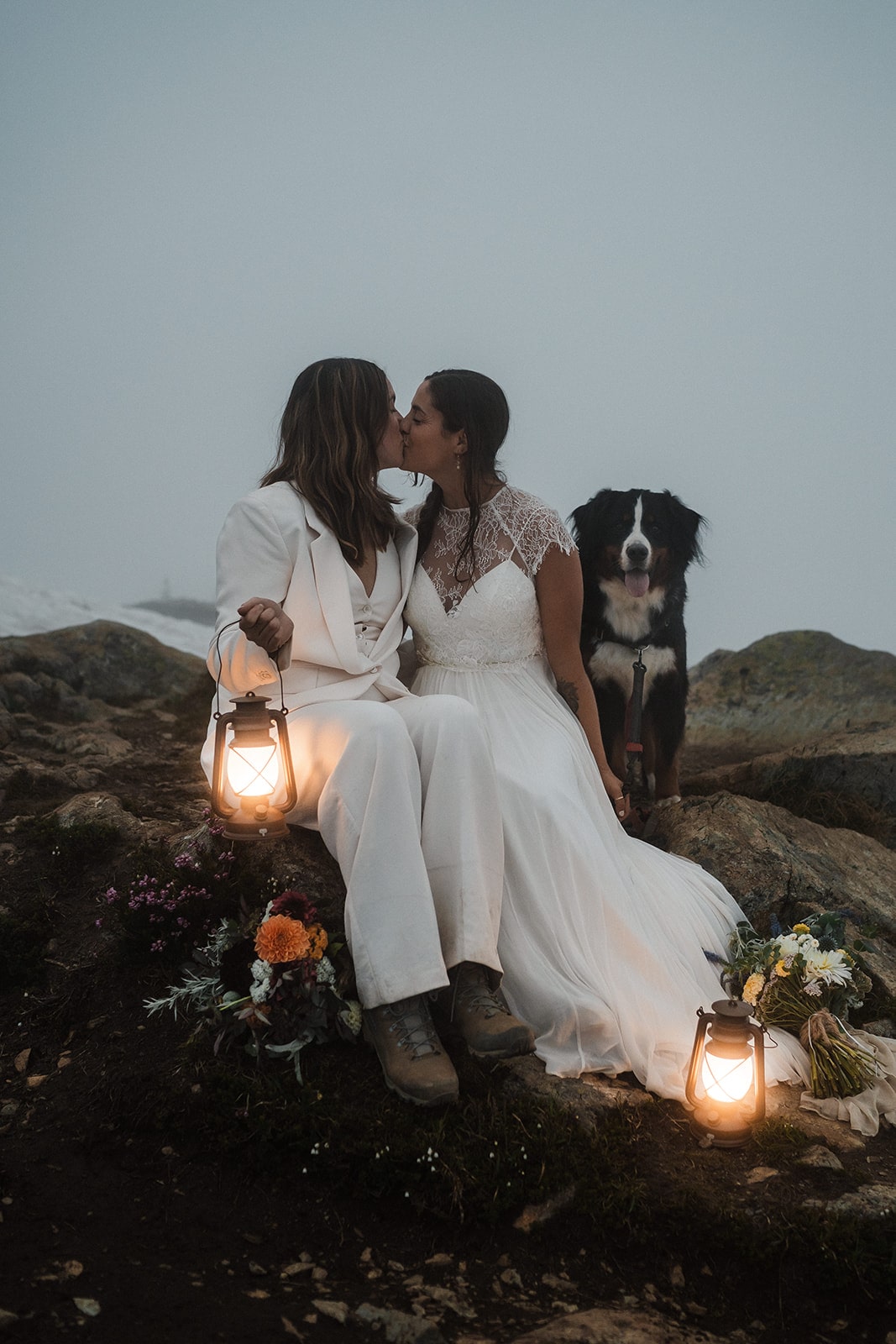 Two brides kiss on a mountain trail next to their dog.