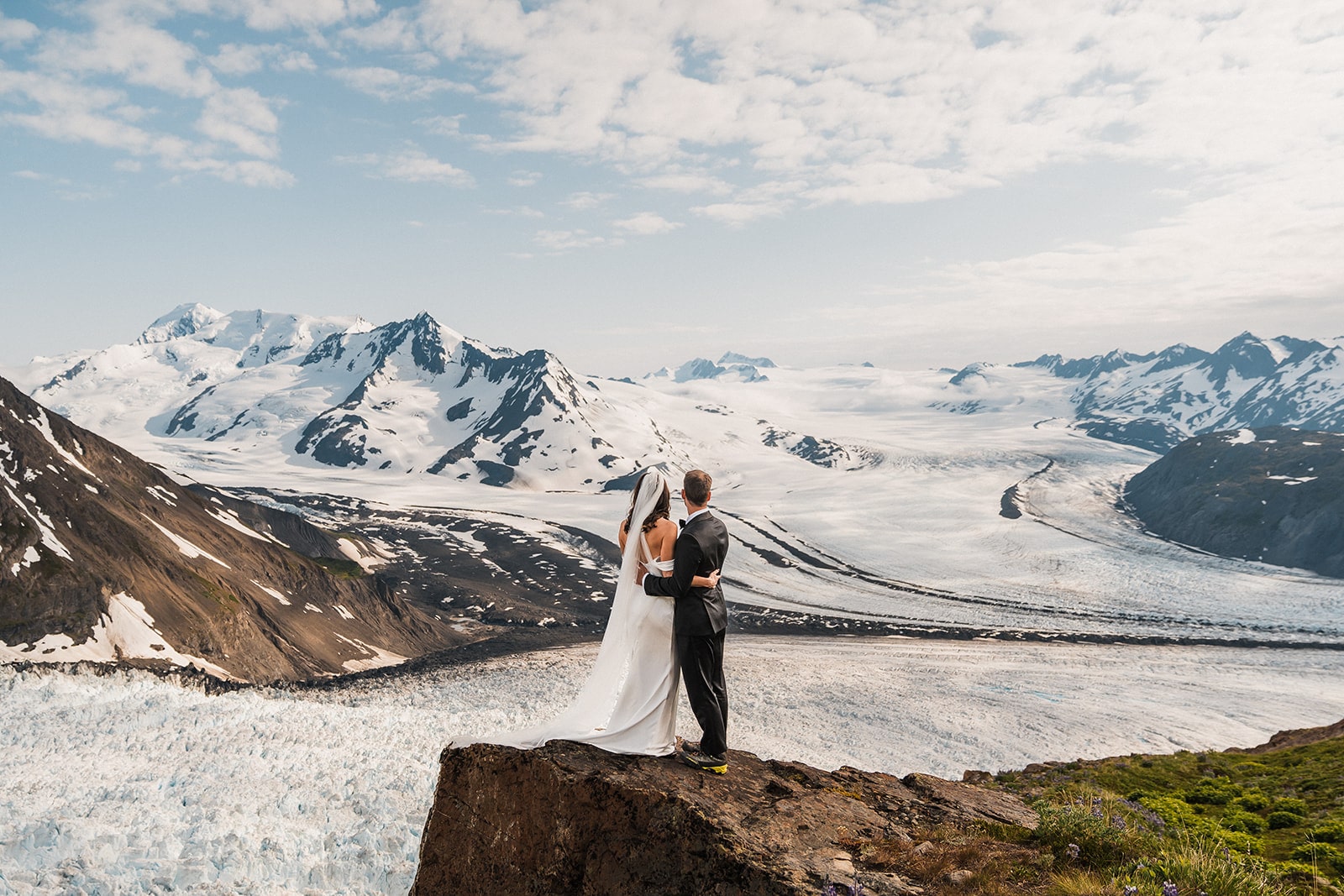 Bride and groom hug on a mountain trail overlooking a glacier during their Alaska elopement. 