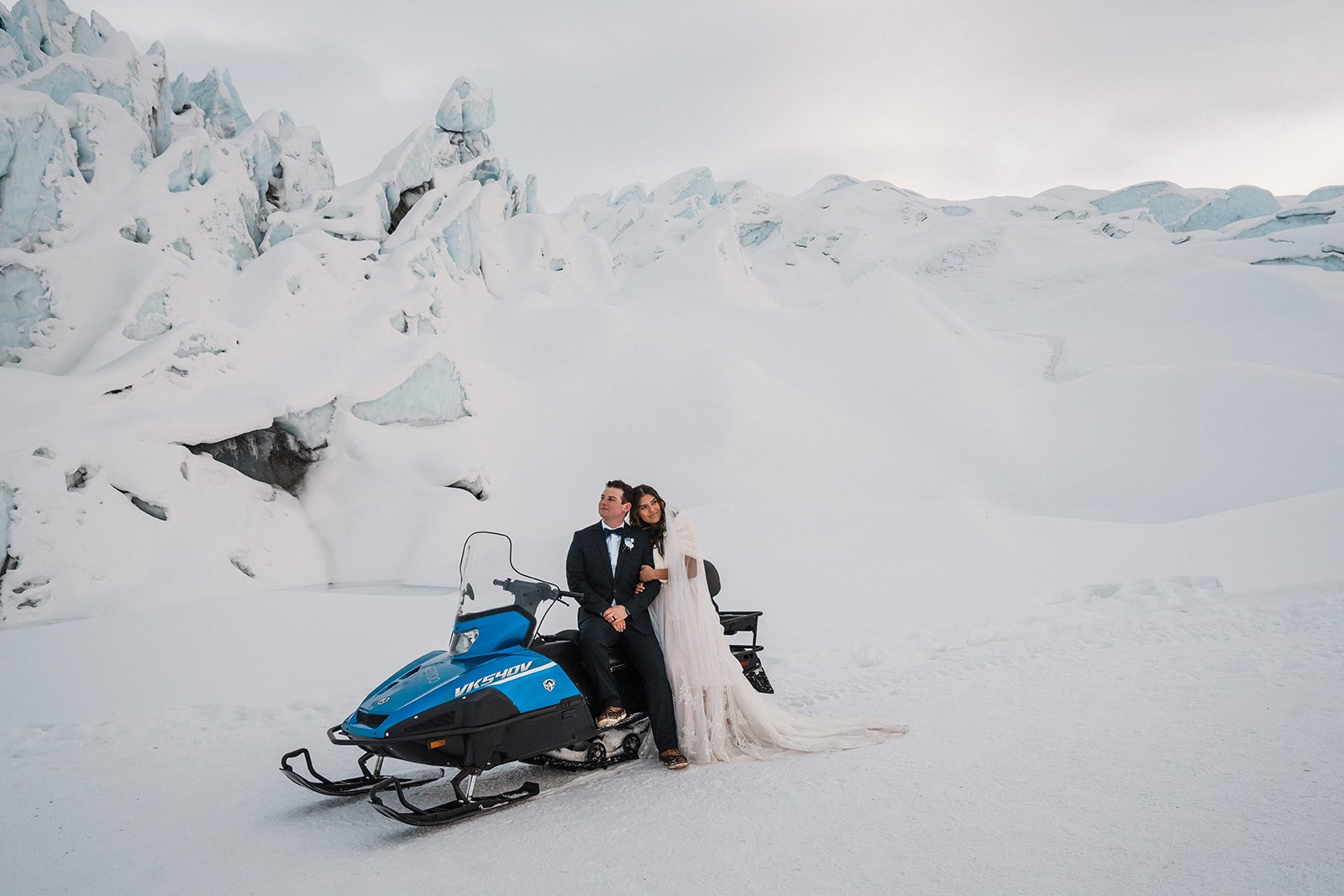Bride and groom sit on a blue snowmobile surrounded by snow after they elope to Alaska. 