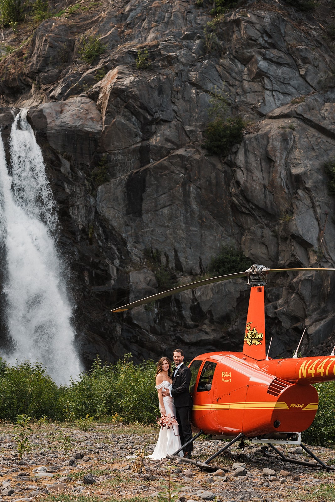 Bride and groom stand under a waterfall next to a red helicopter during their elopement in Alaska.