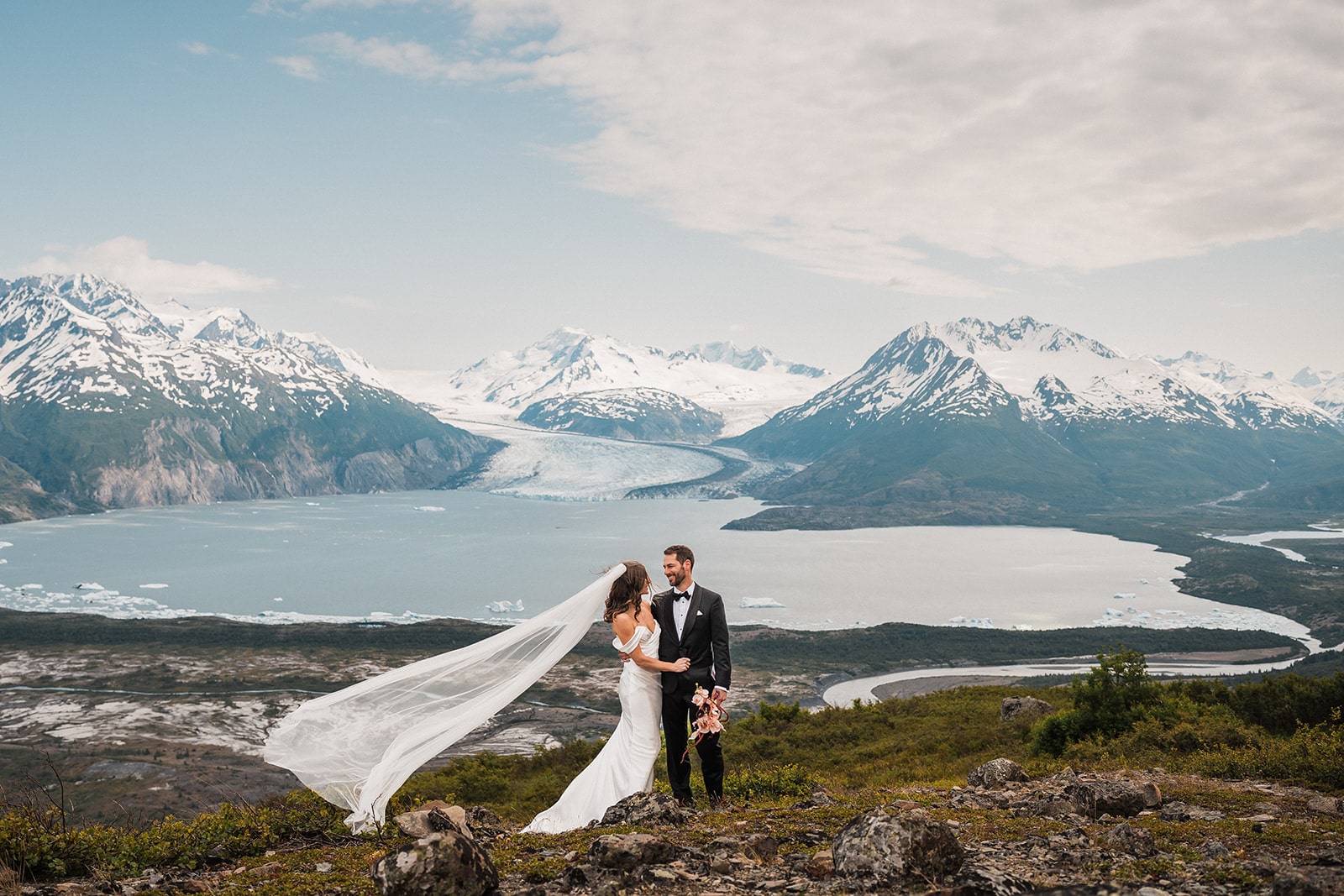 Bride and groom stand beside a mountain lake at their elopement in Alaska. 