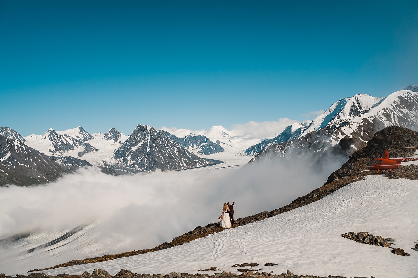 Bride and groom stand on a mountain trail surrounded by snow-covered mountains in Alaska. 