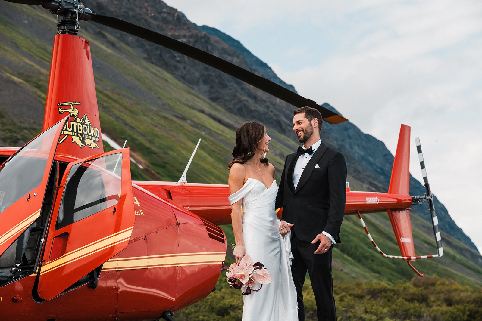 Bride and groom hold hands and laugh next to a red helicopter during their elopement with Outbound Heli Adventures.