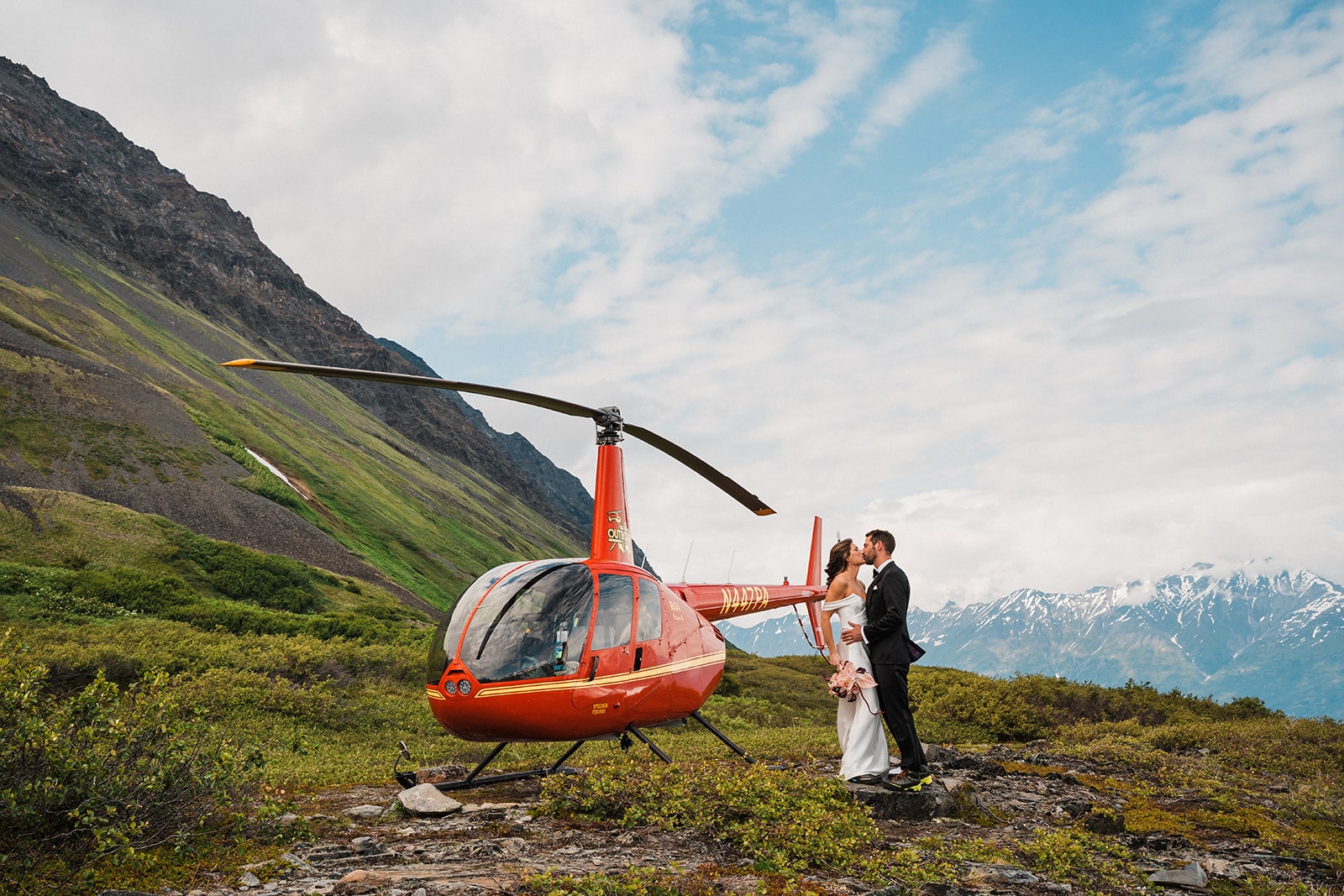 Bride and groom kiss next to a red helicopter on a mountain during their Palmer, Alaska elopement.