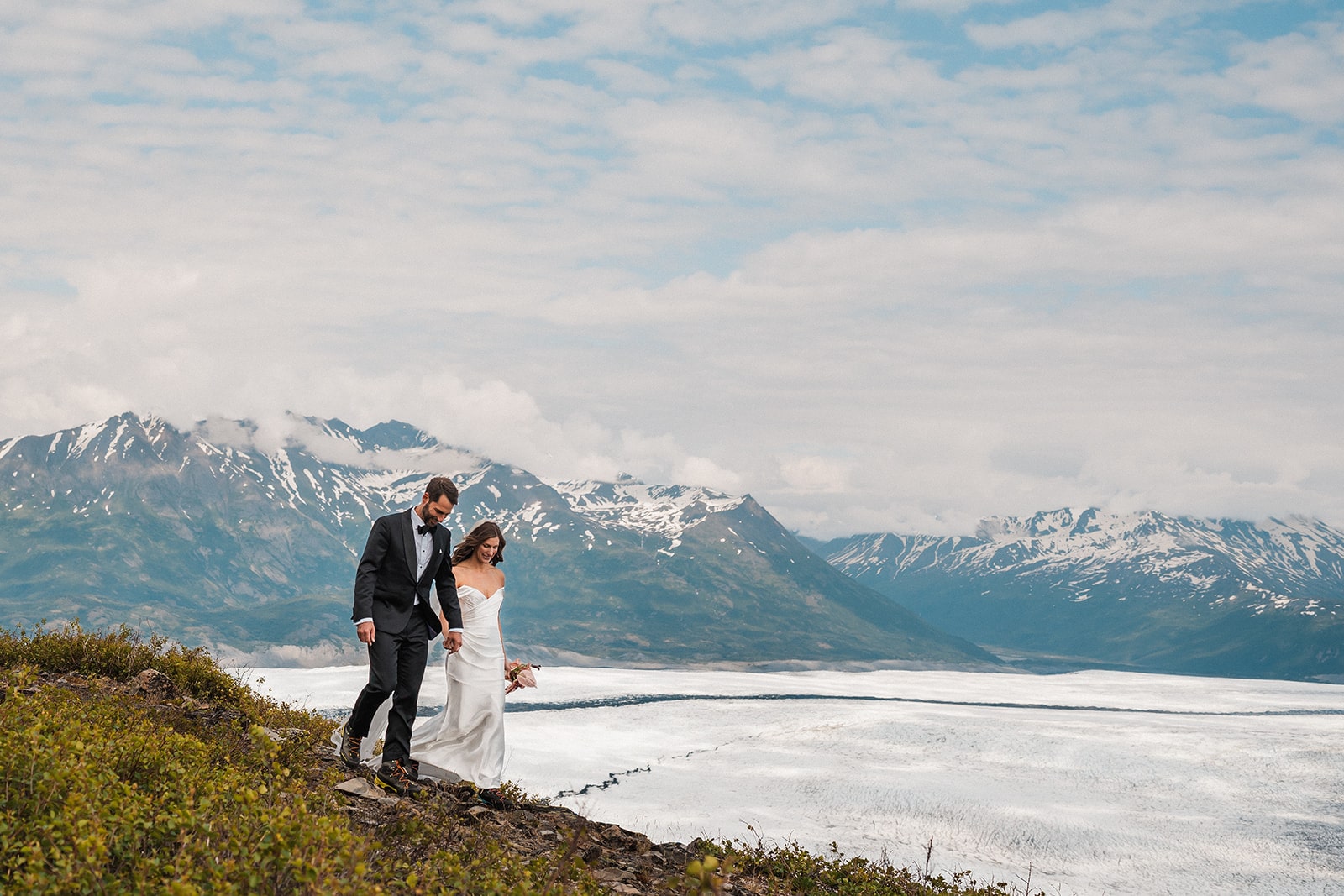 Bride and groom hold hands while walking a mountain during their summer Alaska elopement.