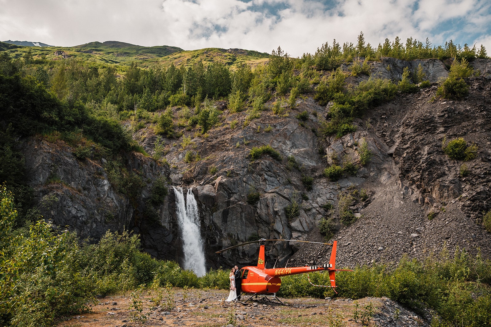 Bride and groom stand next to a red helicopter at the top of a waterfall in Alaska.