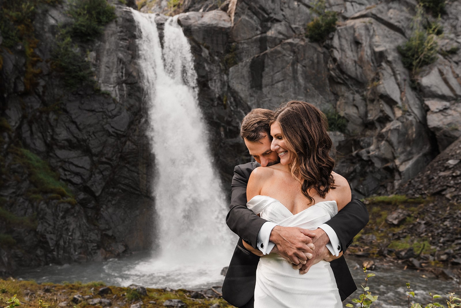 Groom hugs bride from behind as they stand in front of a waterfall at their elopement in Alaska.