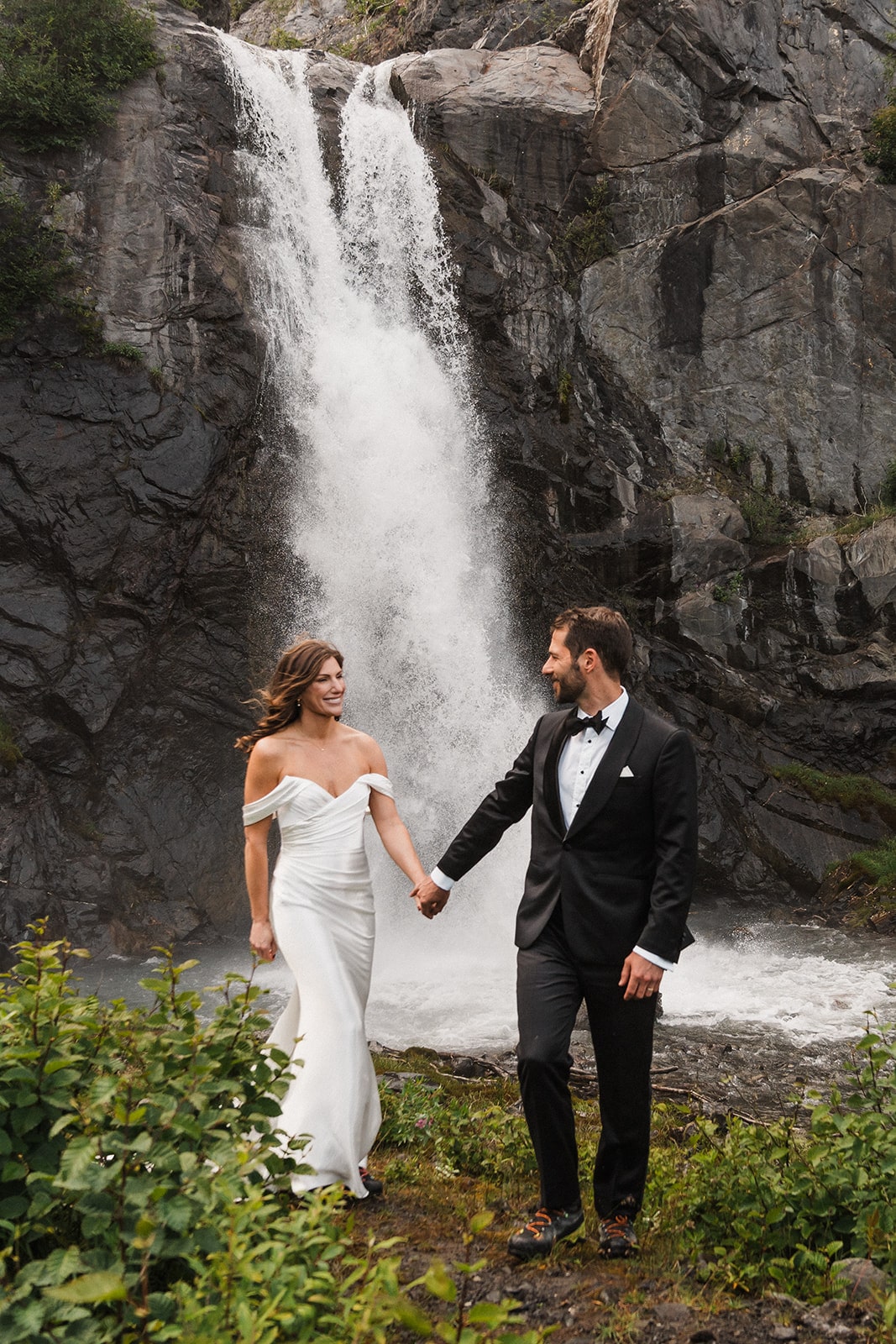 Bride and groom hold hands while walking in front of a waterfall during their summer Alaska elopement.