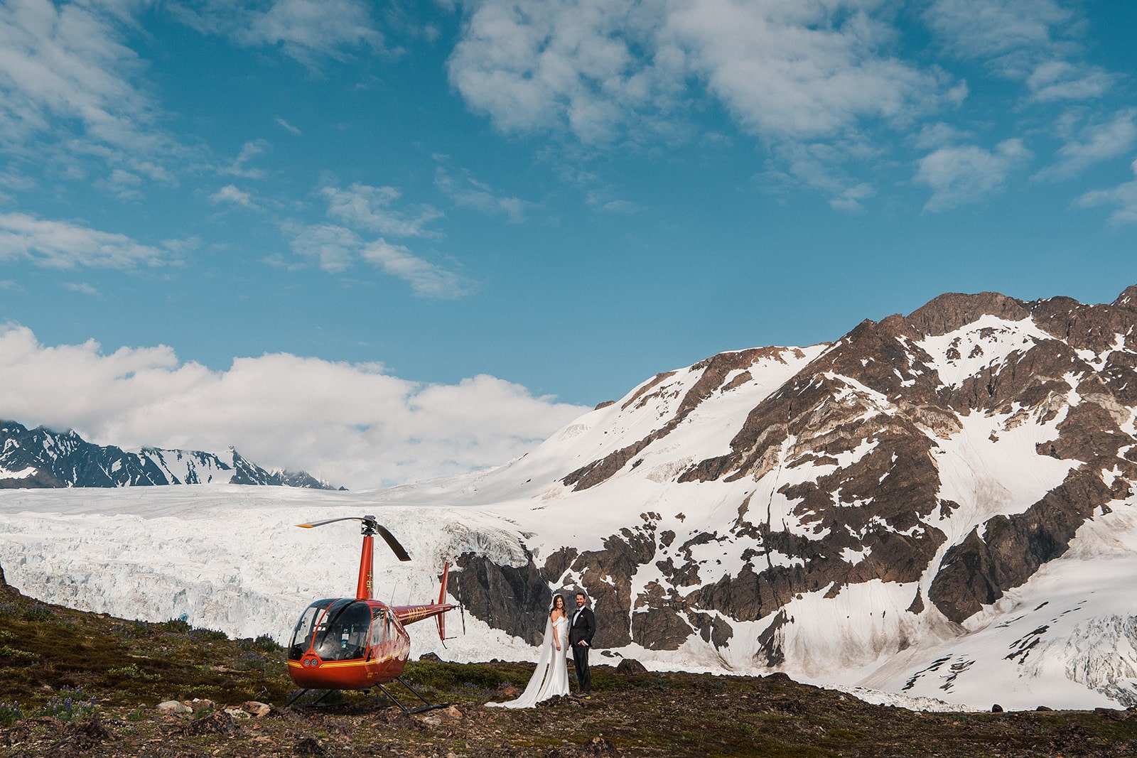 Bride and groom stand on a glacier next to a red helicopter during their elopement with Outbound Heli Adventures.
