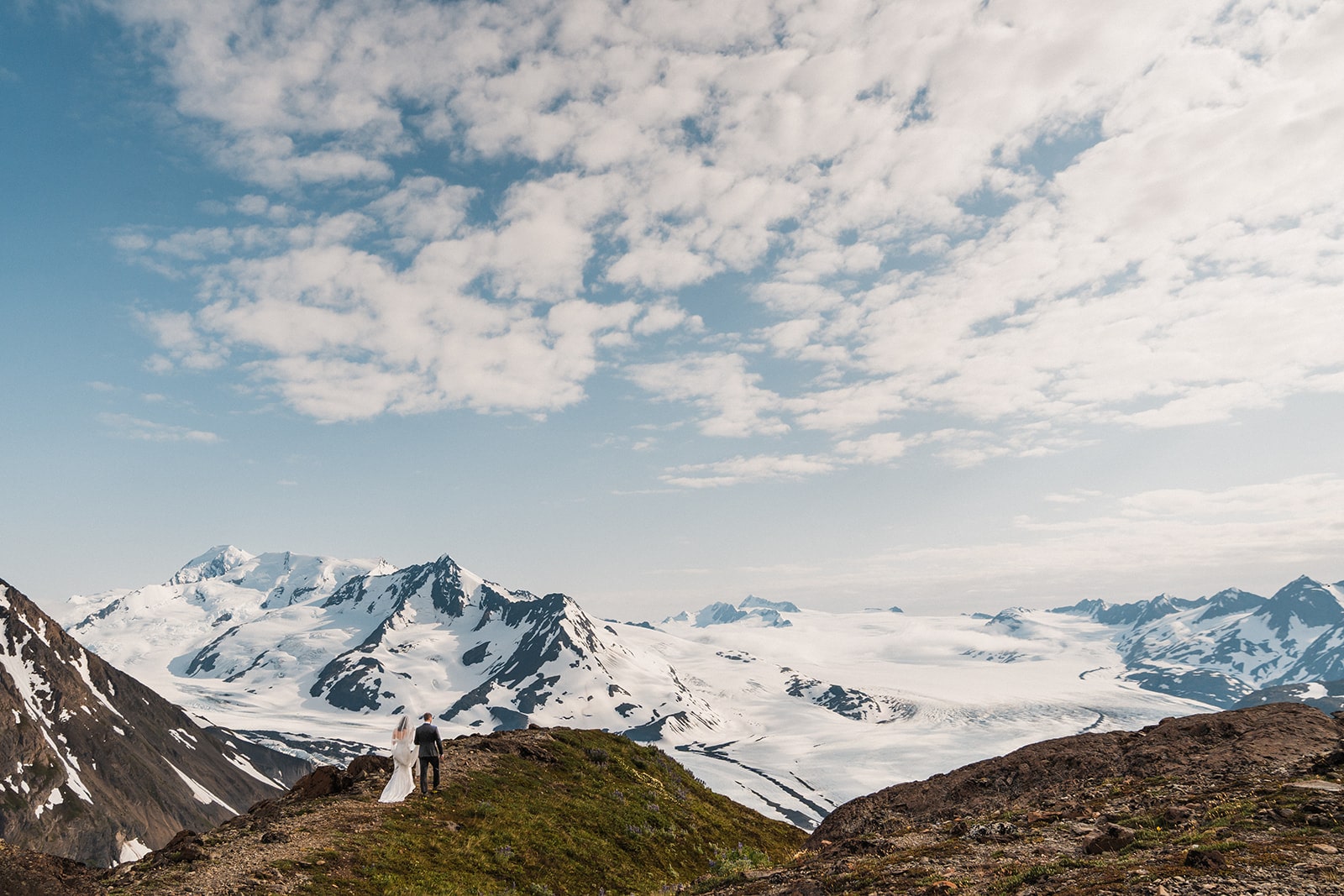 Bride and groom hold hands while walking across a mountain trail with mountain views at their Palmer, Alaska elopement.