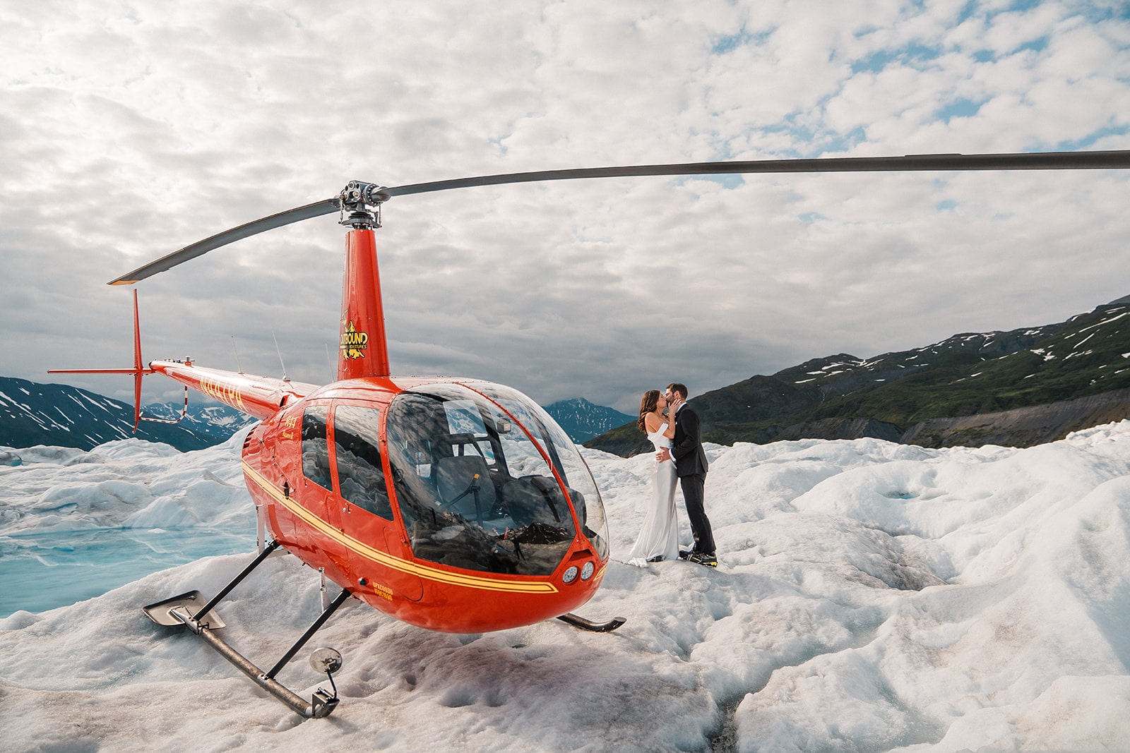 Bride and groom kiss next to a red helicopter on a snowy glacier in Palmer, Alaska.