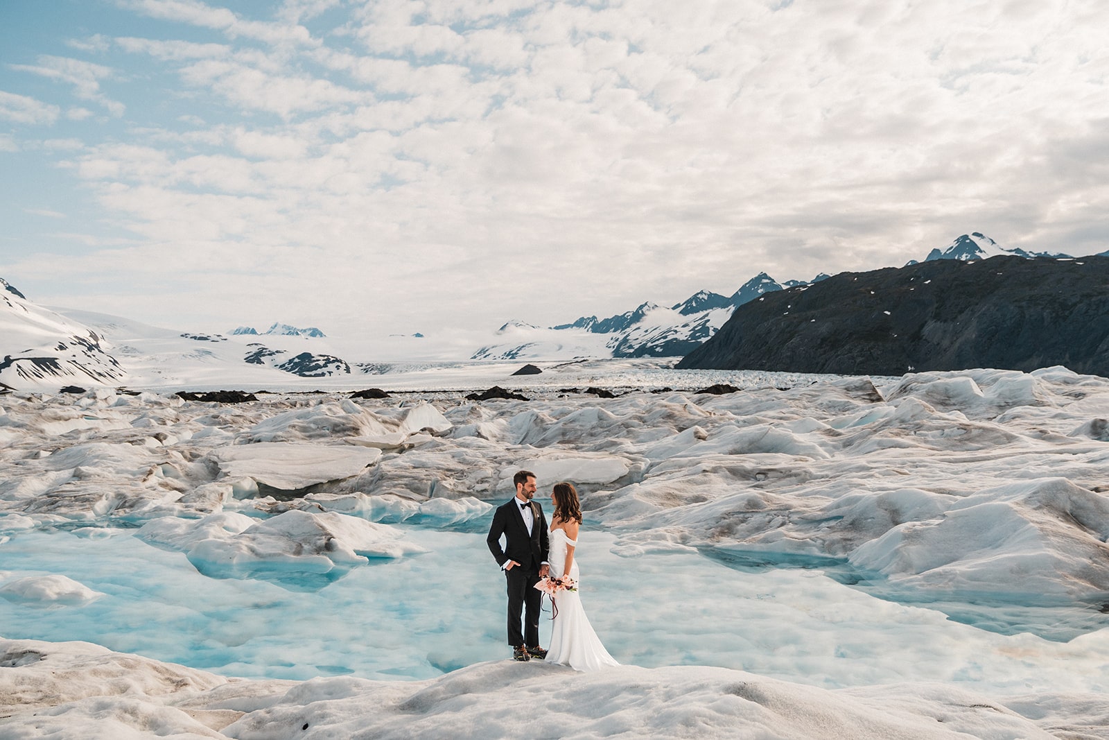 Bride and groom hold hands next to a blue glacier pool during their elopement with Outbound Heli.