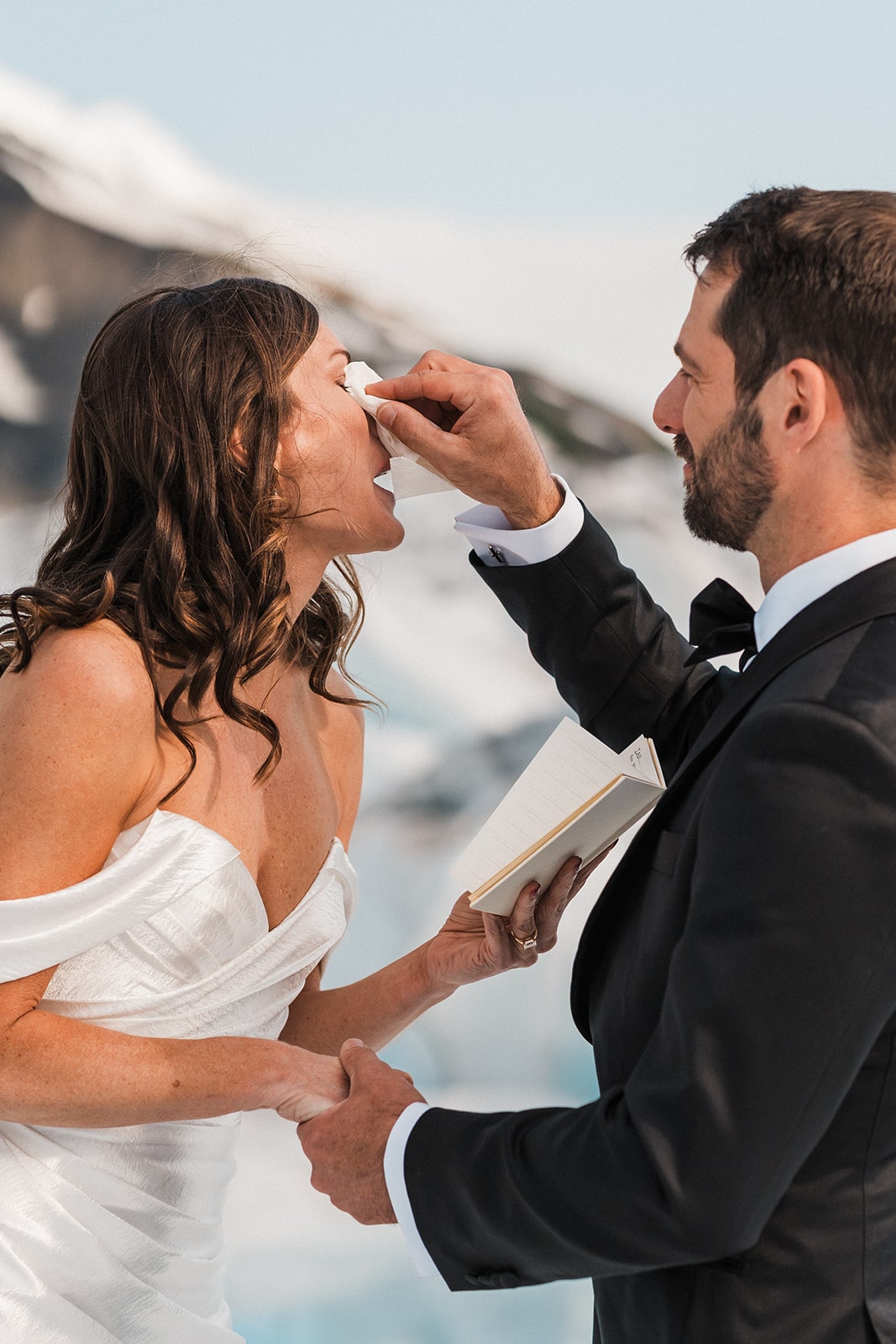 Groom wipes tears from bride's eyes as she reads vows during their Alaska glacier elopement ceremony.