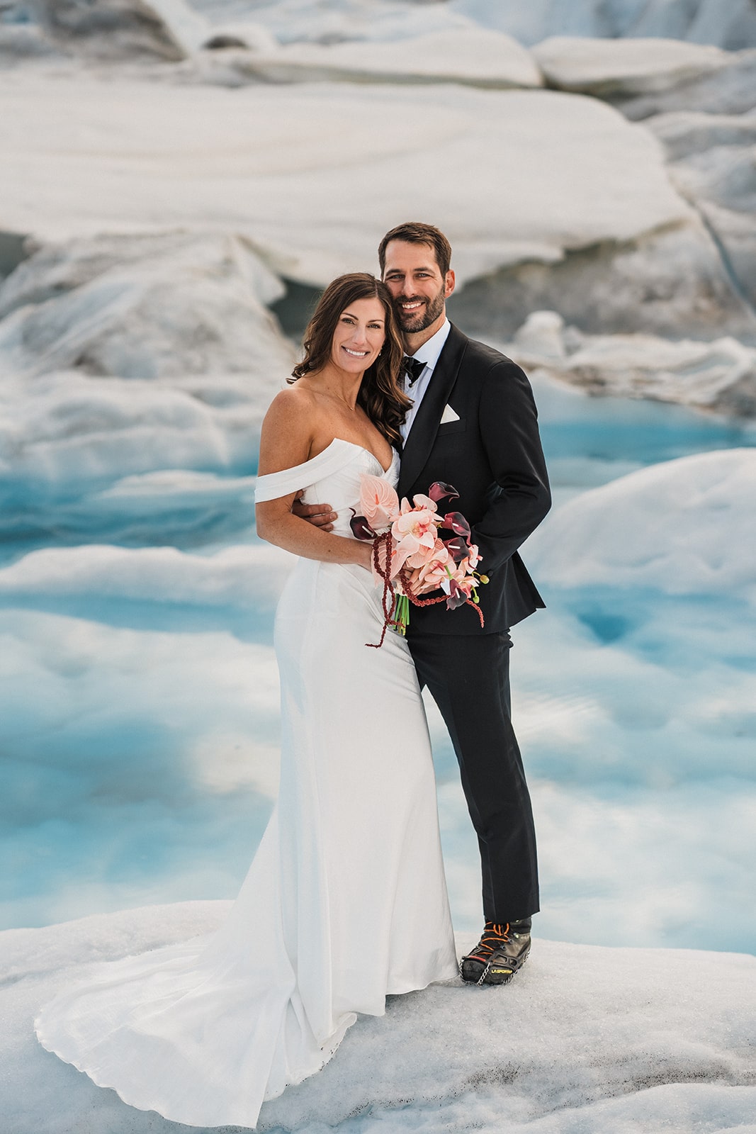 Bride holds pink and burgundy elopement florals during couple portraits with groom on a snowy Alaskan glacier.