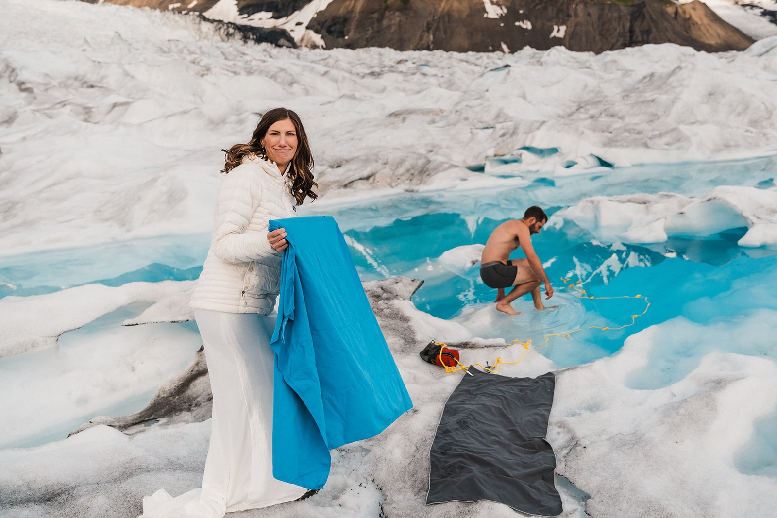 Groom dips into glacier pool while bride stands at the edge holding a blue blanket.