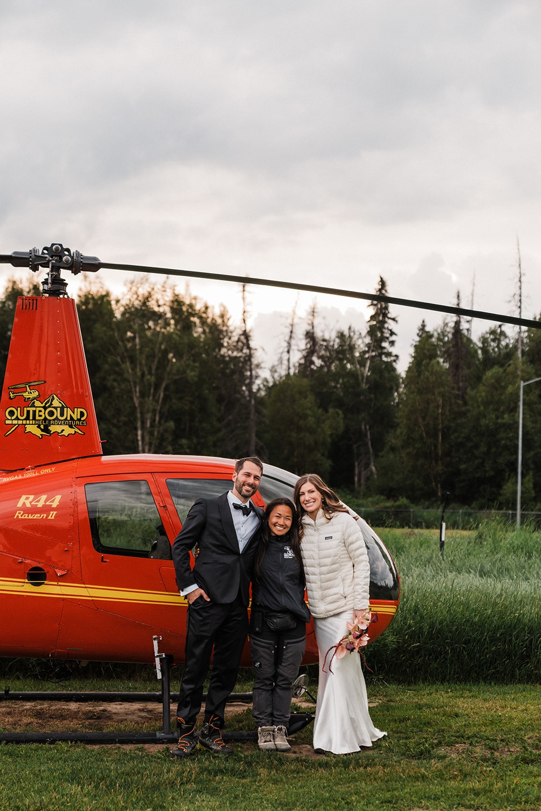 Bride and groom stand next to a red helicopter with their Outbound Heli Adventures pilot.
