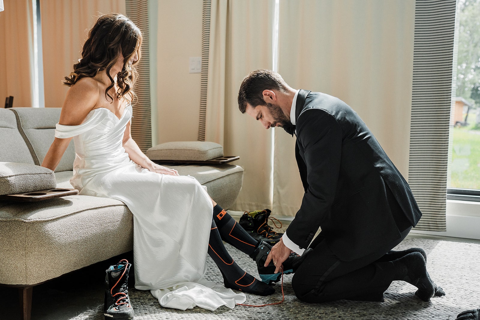 Groom kneels on the floor to help bride put on her hiking shoes.