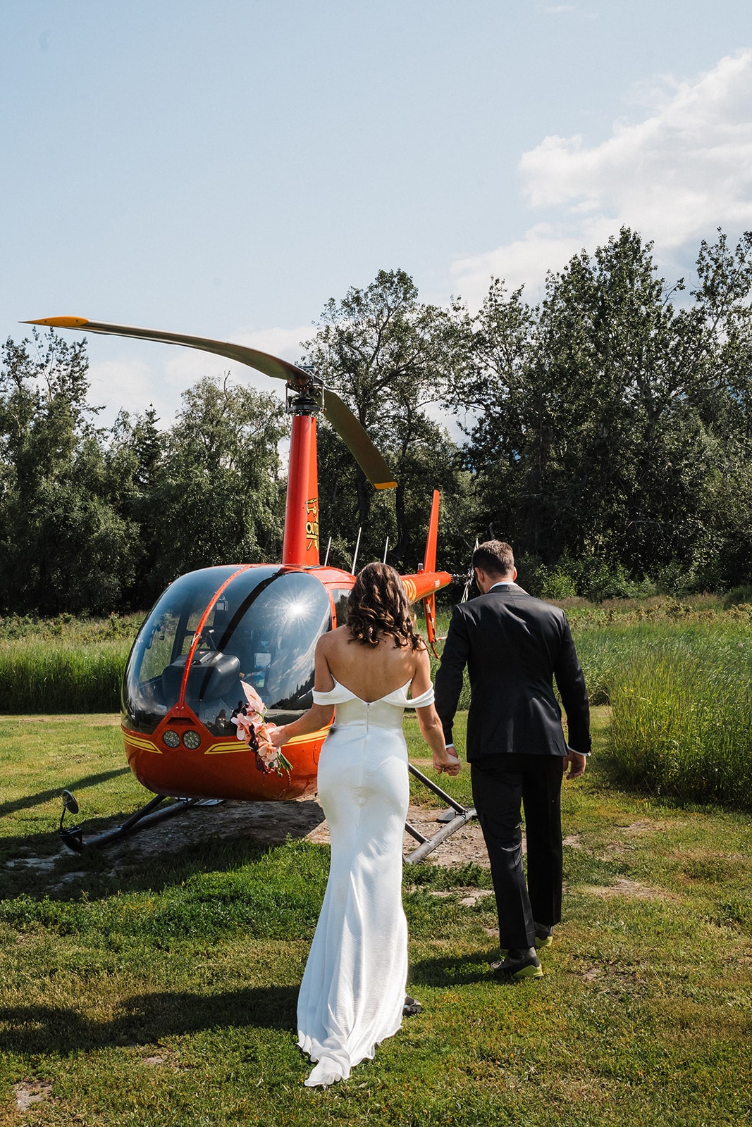 Bride and groom hold hands while walking toward a red helicopter in Palmer, Alaska.