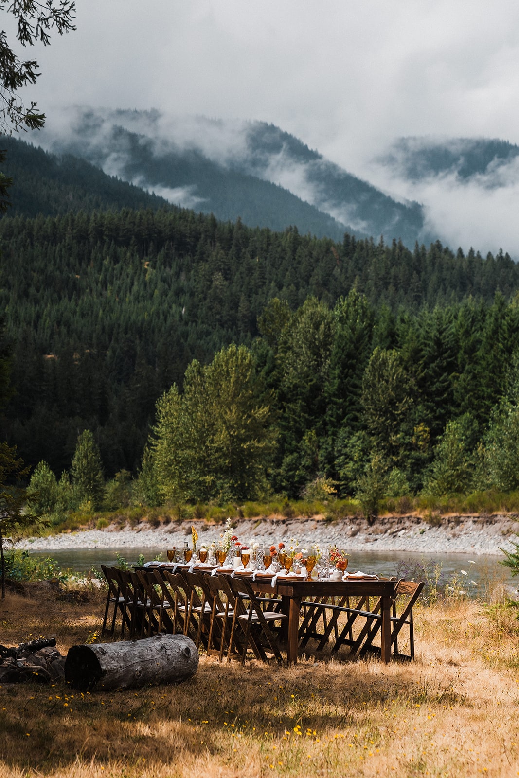 Long wood table decorated with table settings and flowers sits in a field next to a river surrounded by mountains. 