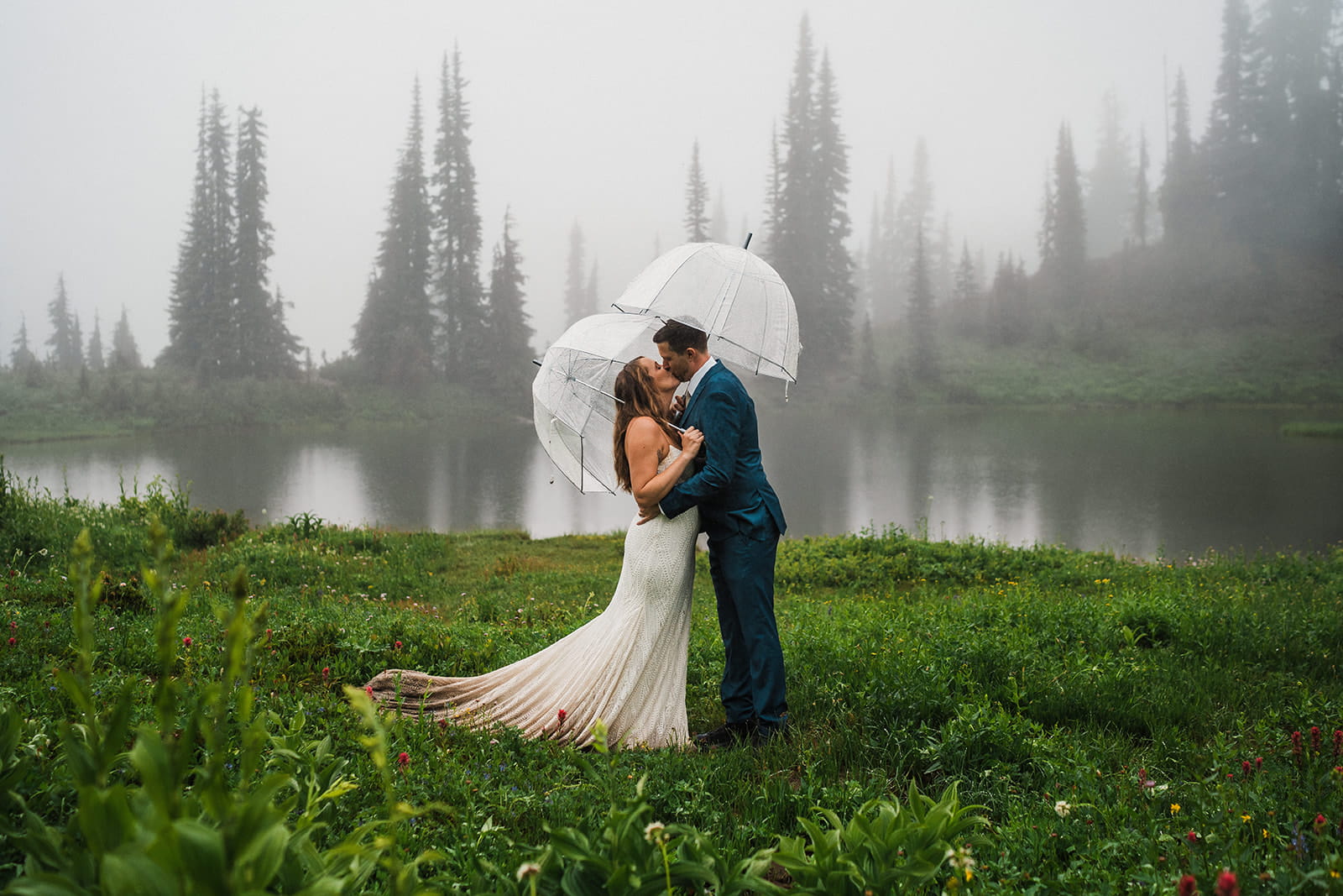 Bride and groom kiss under clear dome umbrella while it rains on their adventure elopement day. 