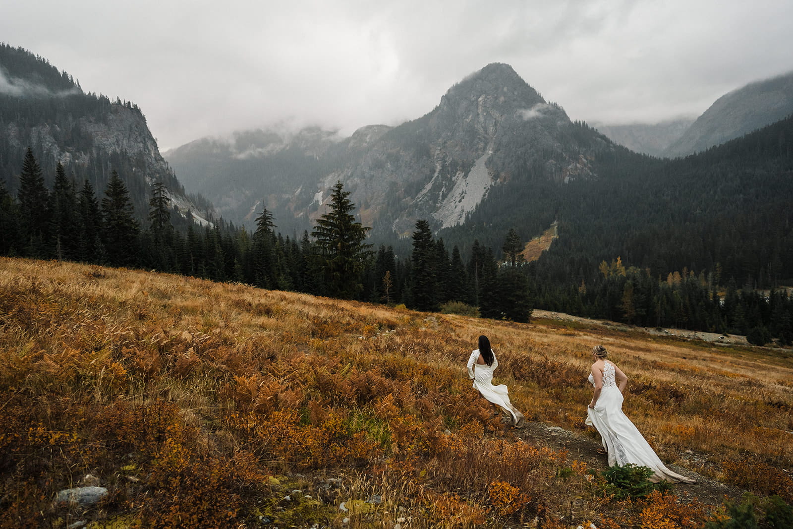 Two brides run through a mountain field during their rainy wedding day photos in Washington. 