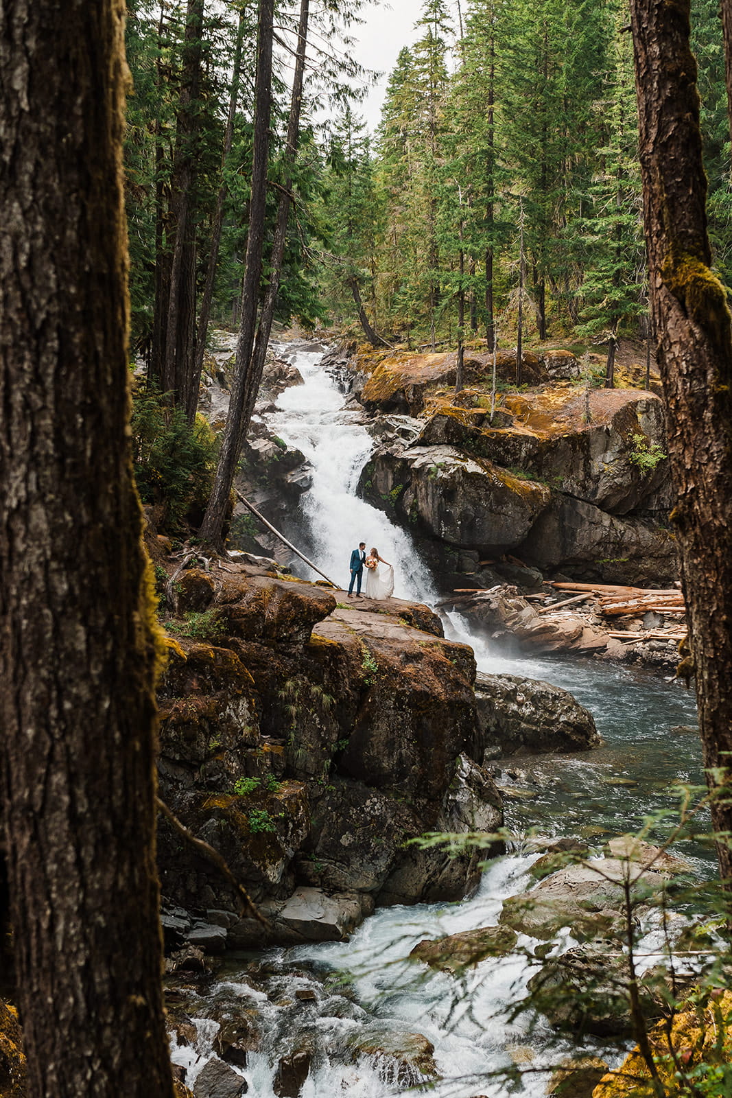 Bride and groom stand on a rocky trail beside a waterfall during their rainy day wedding photos in Washington. 