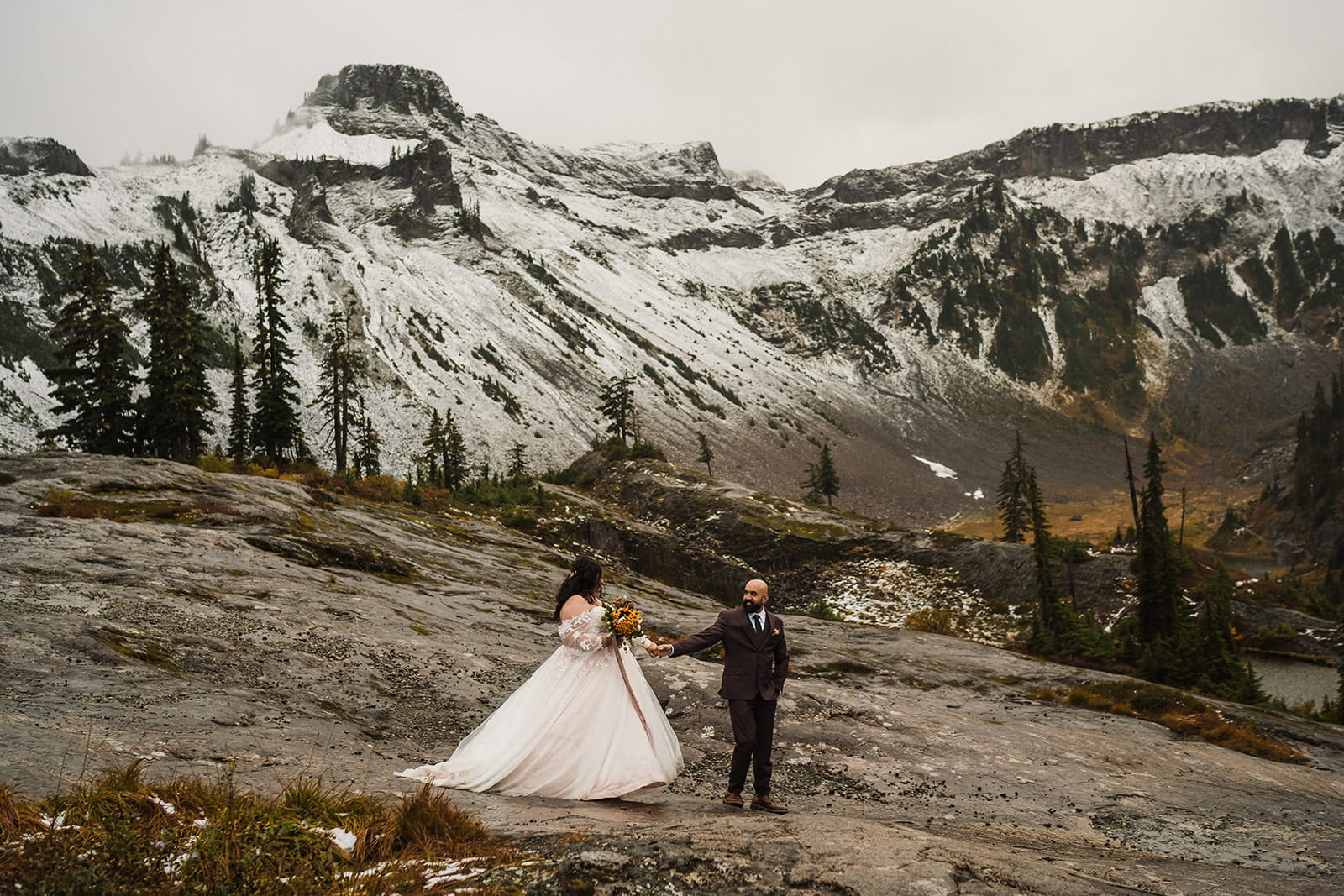 Bride and groom walk across a trail while it snows in the mountains during their elopement wedding. 