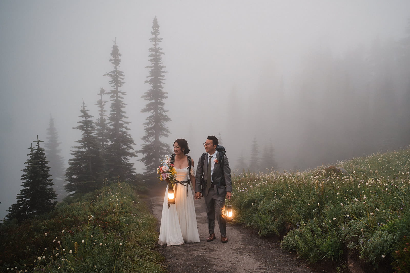 Bride and groom hike a foggy mountain trail with lanterns during their wildflower elopement at Mt Rainier. 