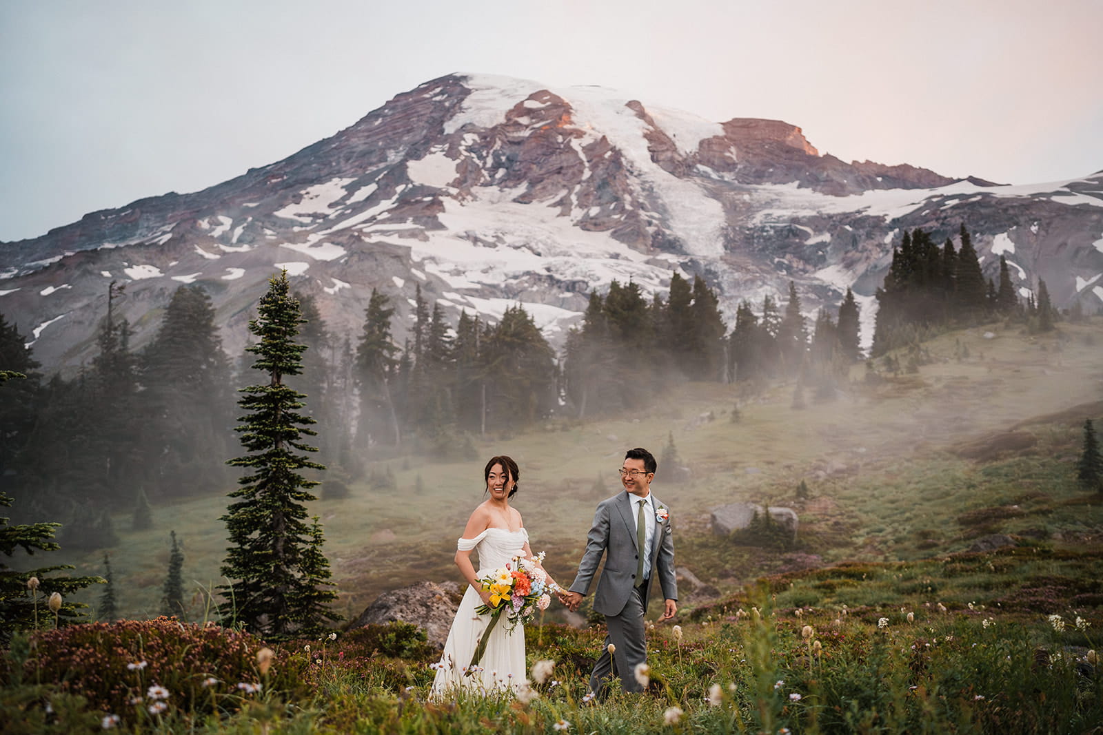 Bride and groom hold hands while walking through a wildflower field in front of Mt Rainier.
