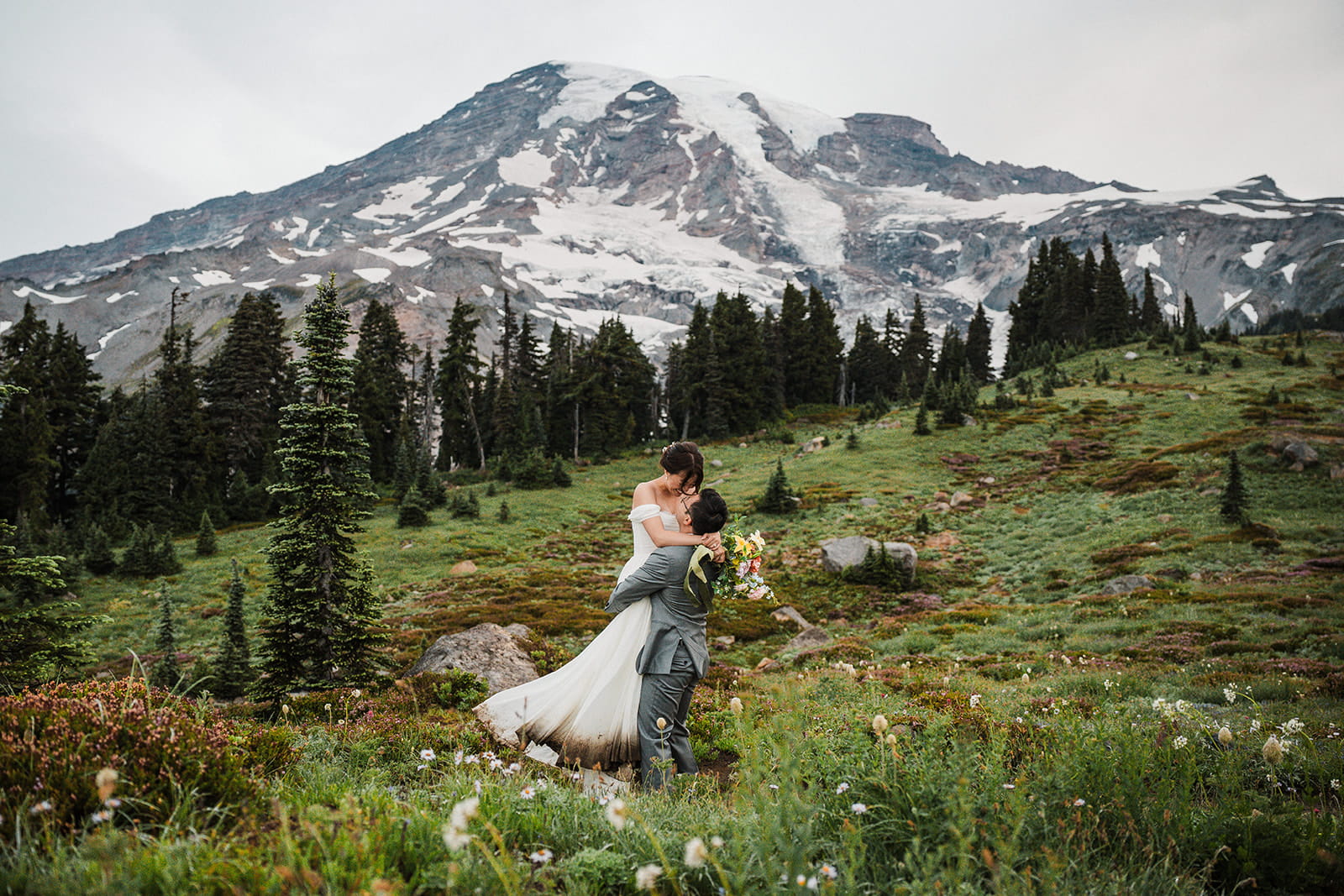 Groom lifts bride up for a kiss on a wildflower mountain trail in Mt Rainier National Park. 