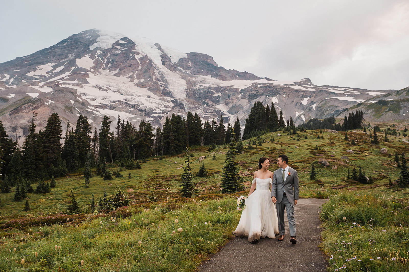 Bride and groom hold hands while walking along a trail in Mt Rainier National Park during their wildflower wedding. 
