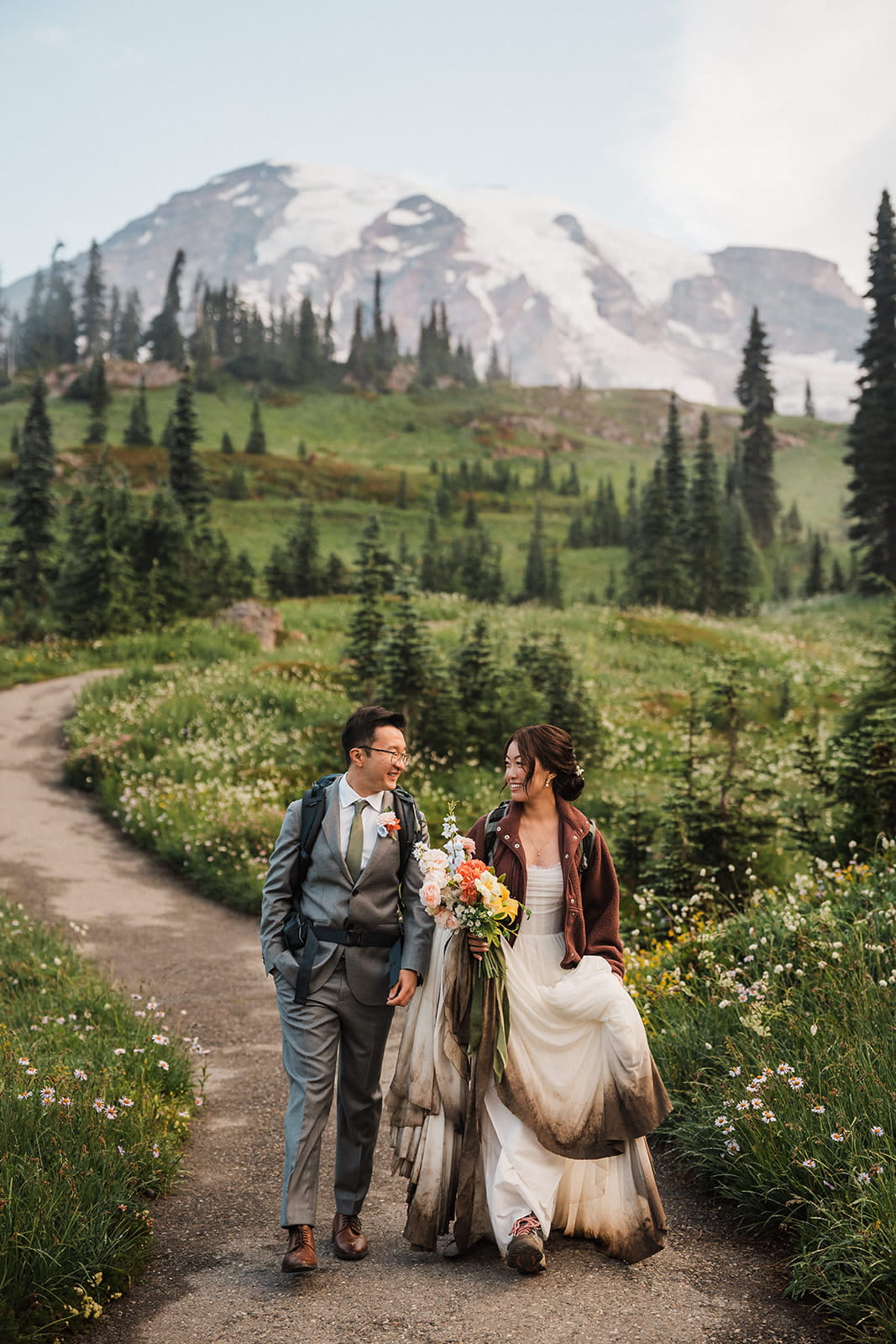 Bride and groom hike in muddy wedding attire on a wildflower trail during their elopement in Mt Rainier National Park. 