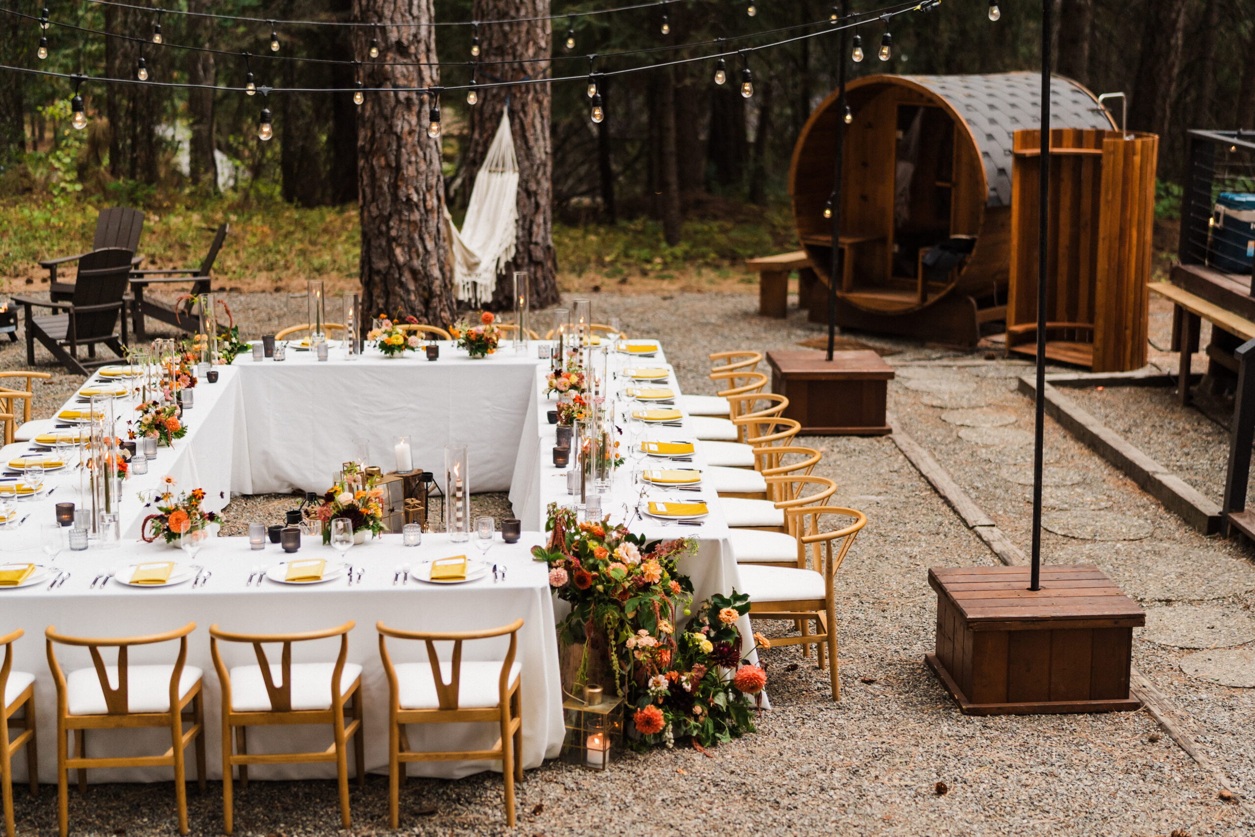 Square reception tables lined with white tablecloths and wood chairs sit in a clearing outside of an A-frame cabin in Washington. 