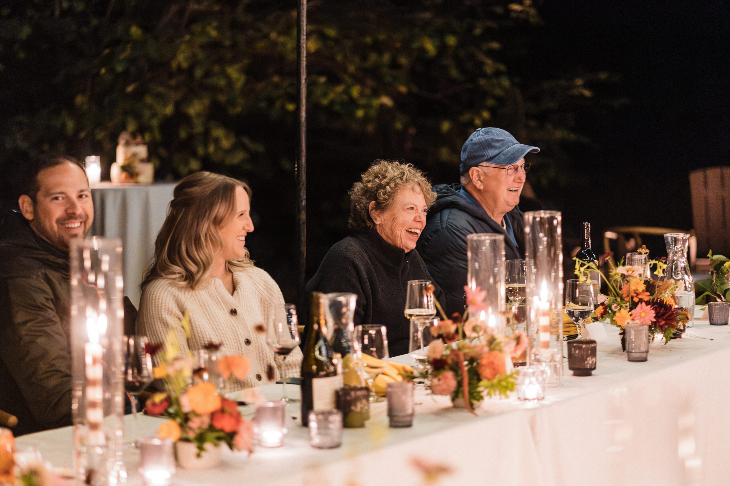 Guests laugh during toasts at an outdoor candlelight wedding reception dinner.