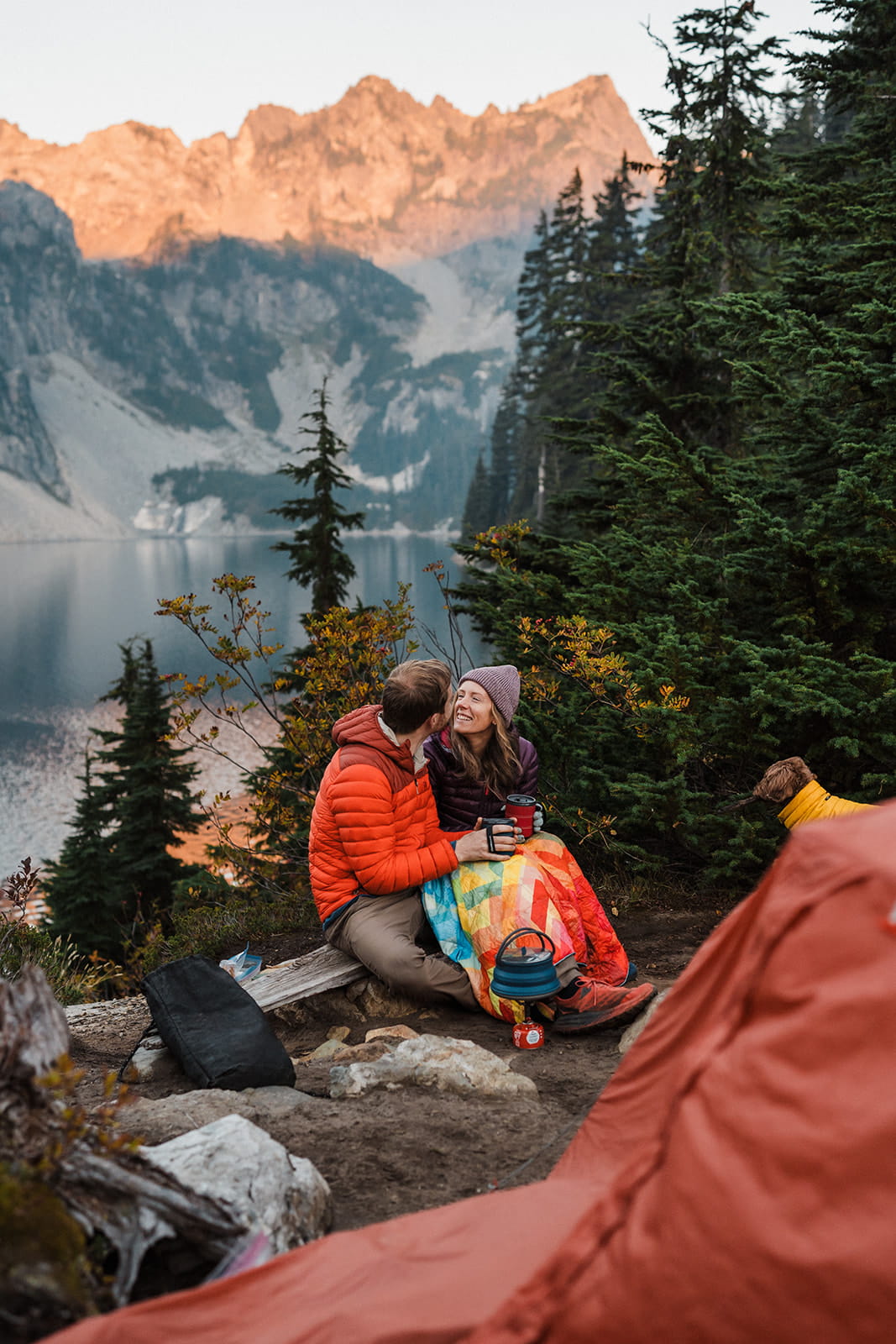 Couple sits on a log drinking coffee at sunrise during their backpacking elopement. 