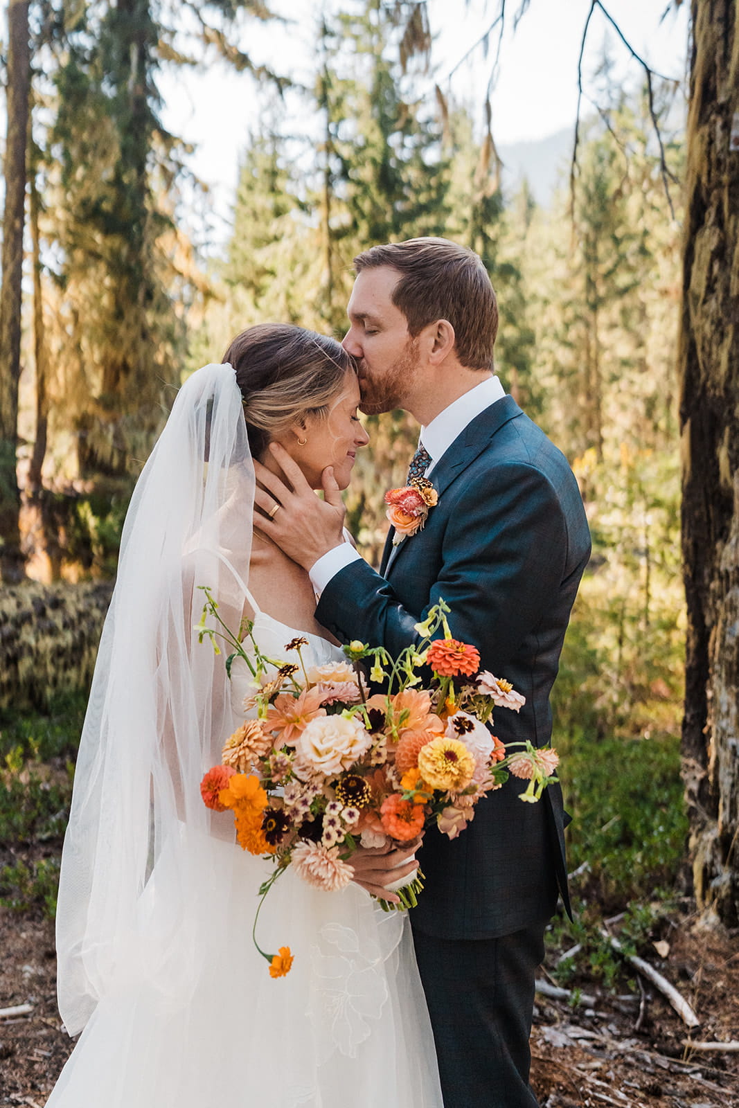 Groom kisses bride on the forehead during their forest wedding photos. 