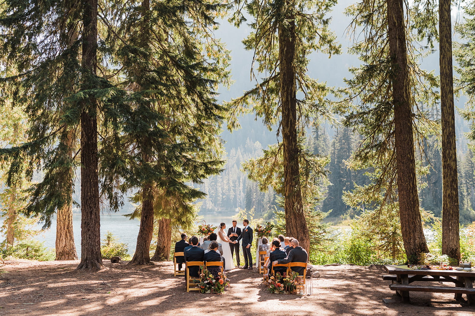 Guests sit on wood chairs in a forest clearing by Cooper Lake. 