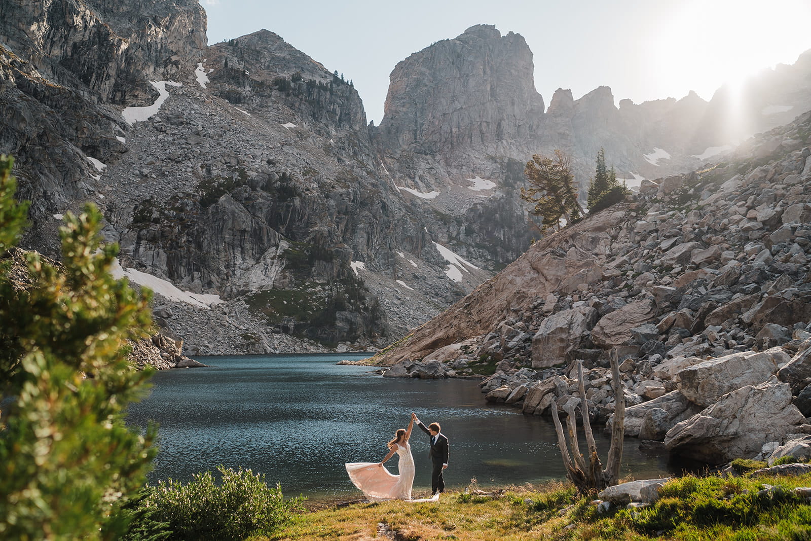 Couple in wedding attire dance by an alpine lake surrounded by mountain peaks.