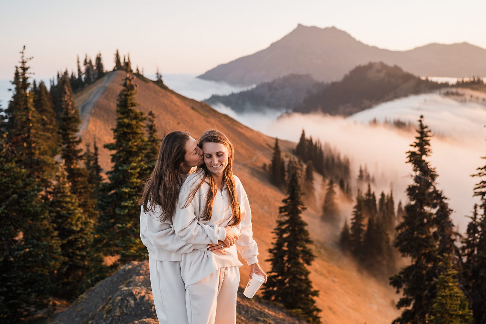 Two women in sweatsuits hug on a mountain at sunrise in Olympic National Park.