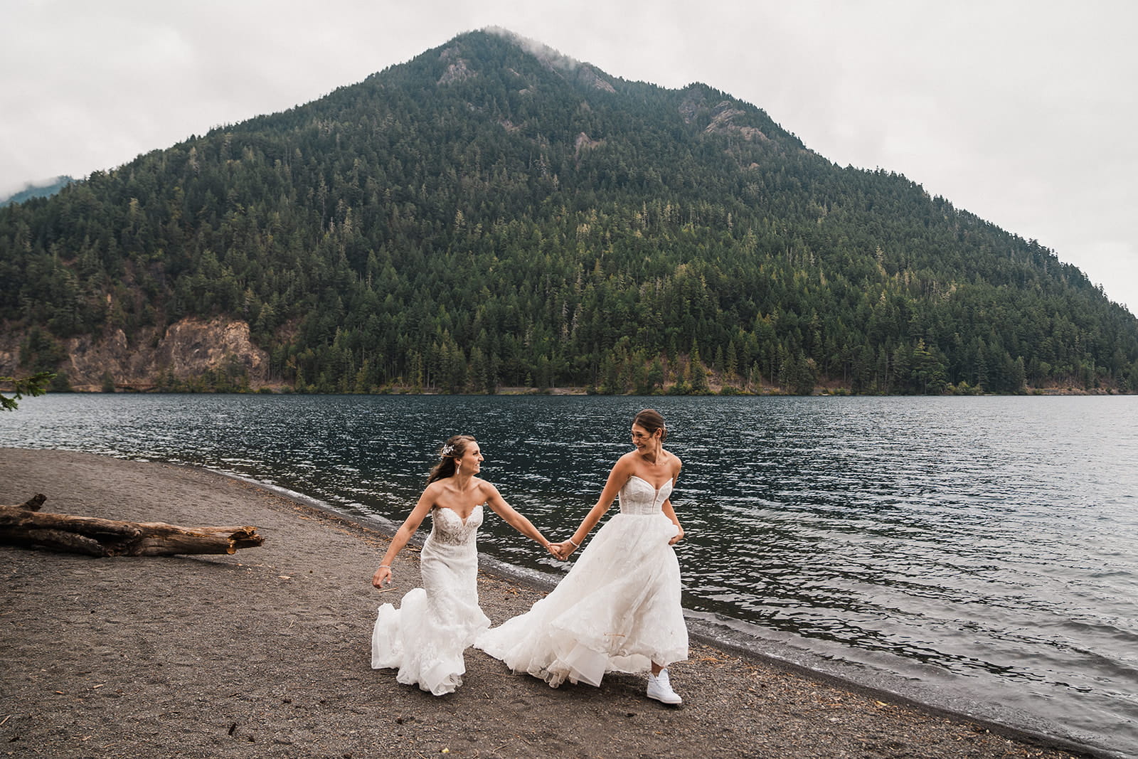 Two brides in white wedding dresses run across the beach at Lake Crescent.